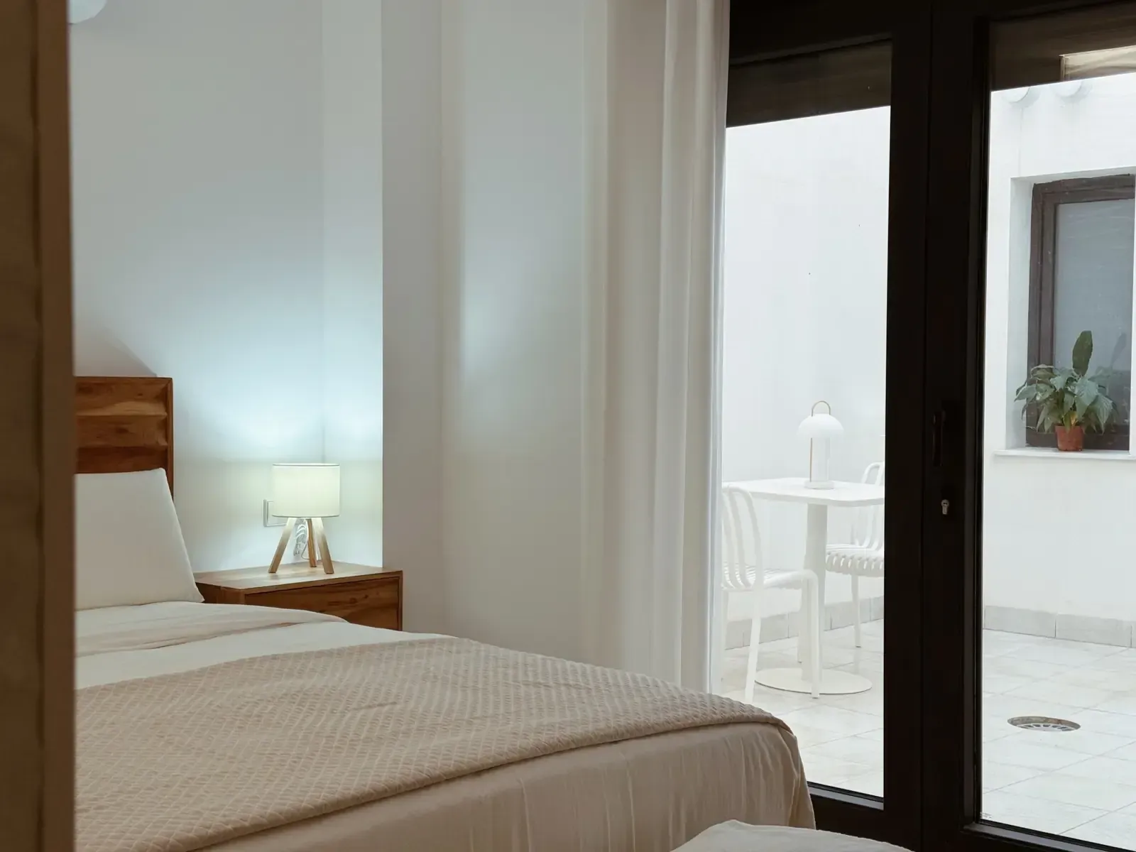 Un dormitorio con cama y mesita de noche de madera, con acceso a un patio blanco con una mesa pequeña y sillas.