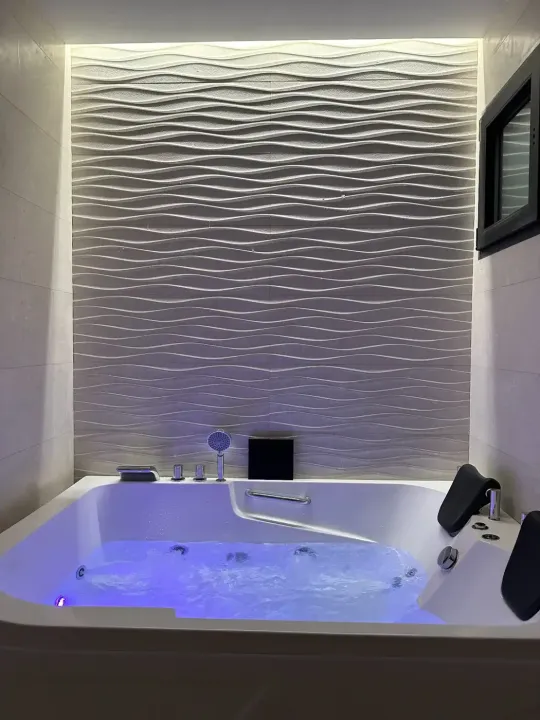 Un jacuzzi blanco con agua azul brillante, situado contra una pared texturizada con diseño de olas e iluminado con luz tenue.