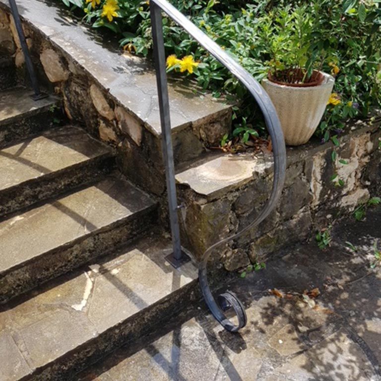 Des marches en pierre avec une rampe en métal jouxtent une jardinière ensoleillée.