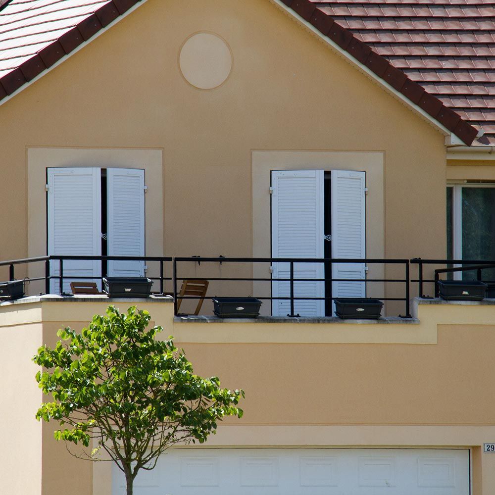 Façade de maison beige avec balcon, volets blancs, toit de tuiles rouges et un petit arbre devant.