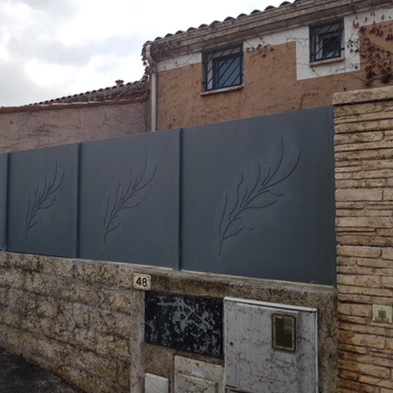 Mur en pierre et clôture grise préservant l'intimité devant une maison, ornée de motifs de feuilles et du numéro 48 visible sur le portail.