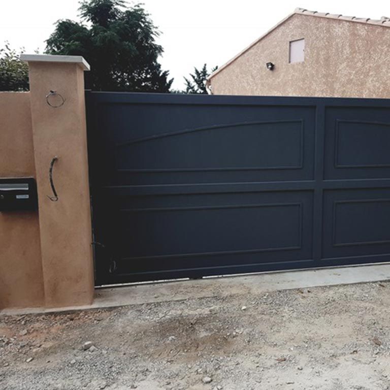 Portail d'allée en métal noir jouxtant un mur en stuc beige et une allée en gravier.