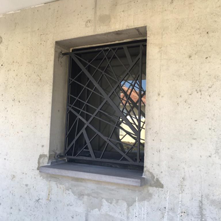 Une fenêtre à barreaux encastrée dans un mur beige délavé, avec un petit appui et un plâtre qui s'écaille.
