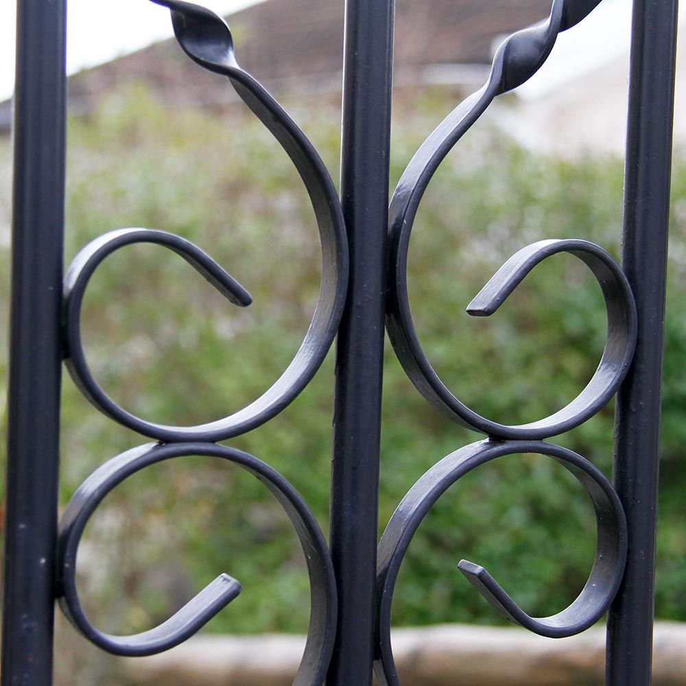 Gros plan sur un portail en fer forgé noir orné de volutes décoratives et de verdure floue en arrière-plan.
