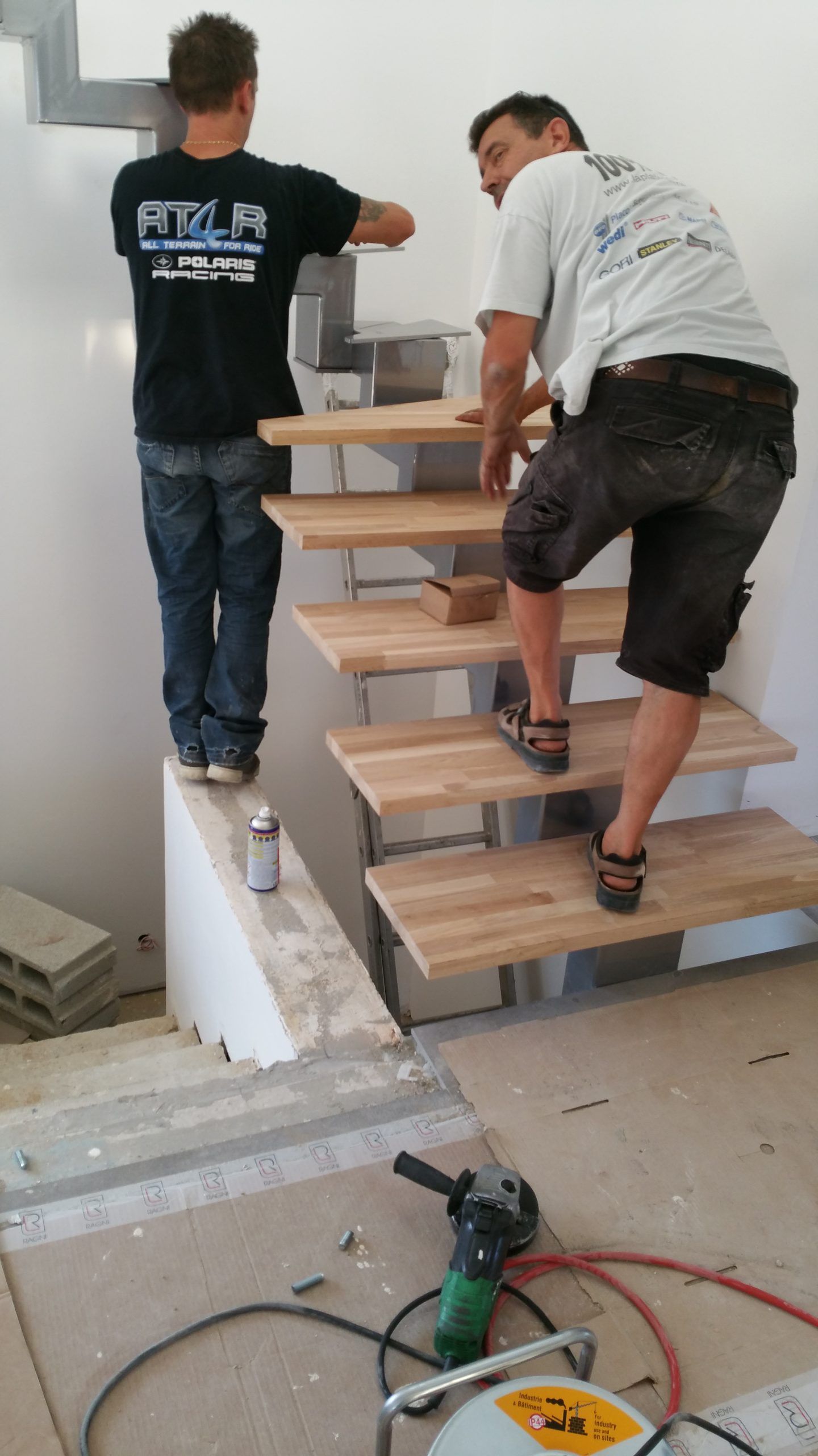 Deux ouvriers transportent des marches en bois le long d'une structure d'escalier blanche à l'intérieur.