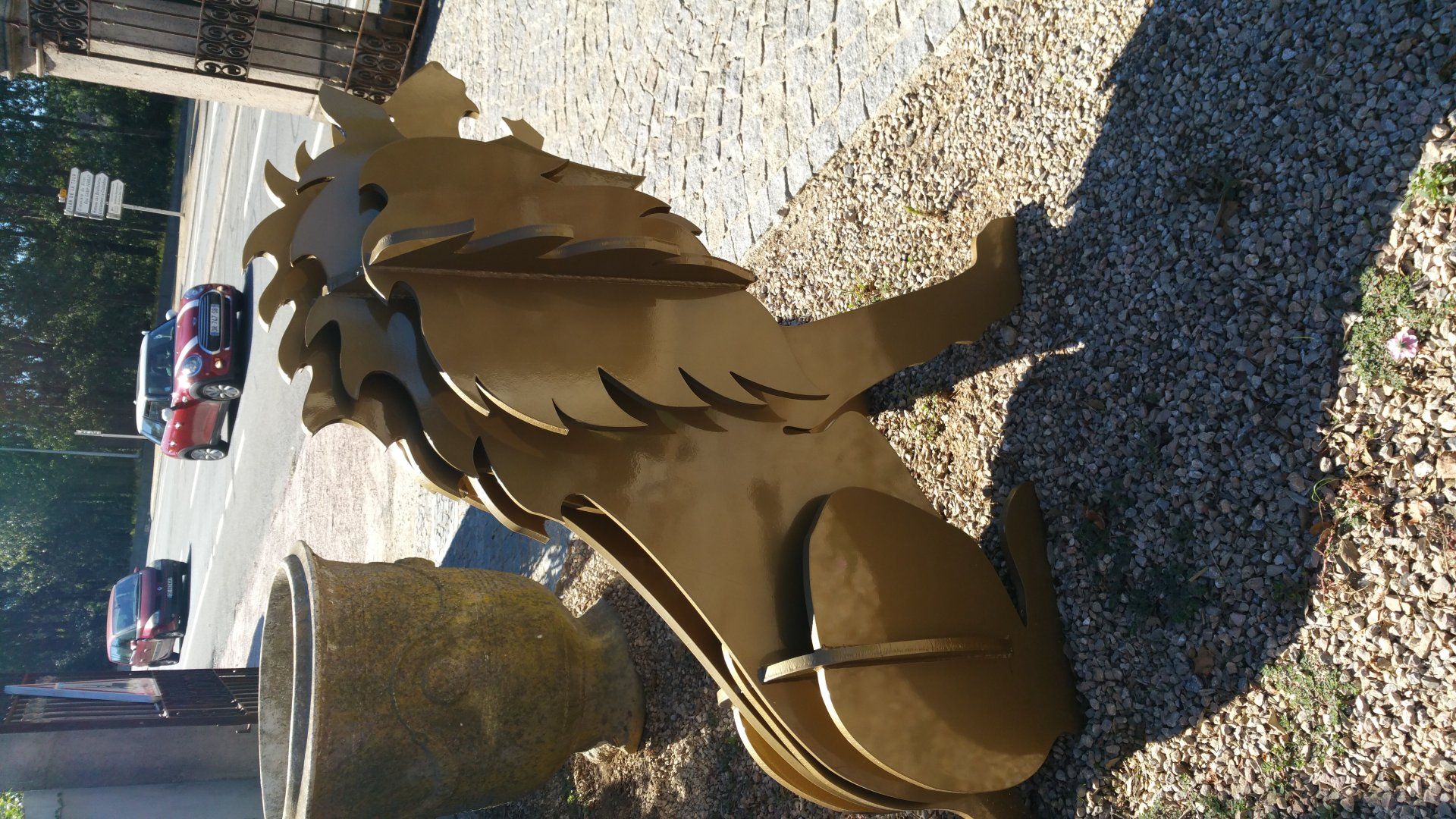 Sculpture d'une créature assise ressemblant à un dragon, au bord d'un chemin de gravier, avec des voitures garées en arrière-plan.