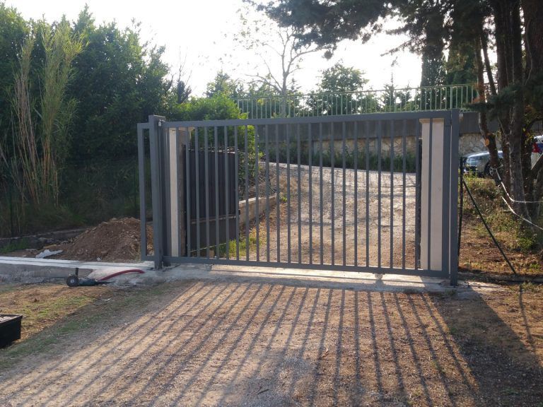 Portail métallique à l'entrée d'une allée, projetant des ombres sur le chemin de gravier, bordé d'arbres et d'une clôture.