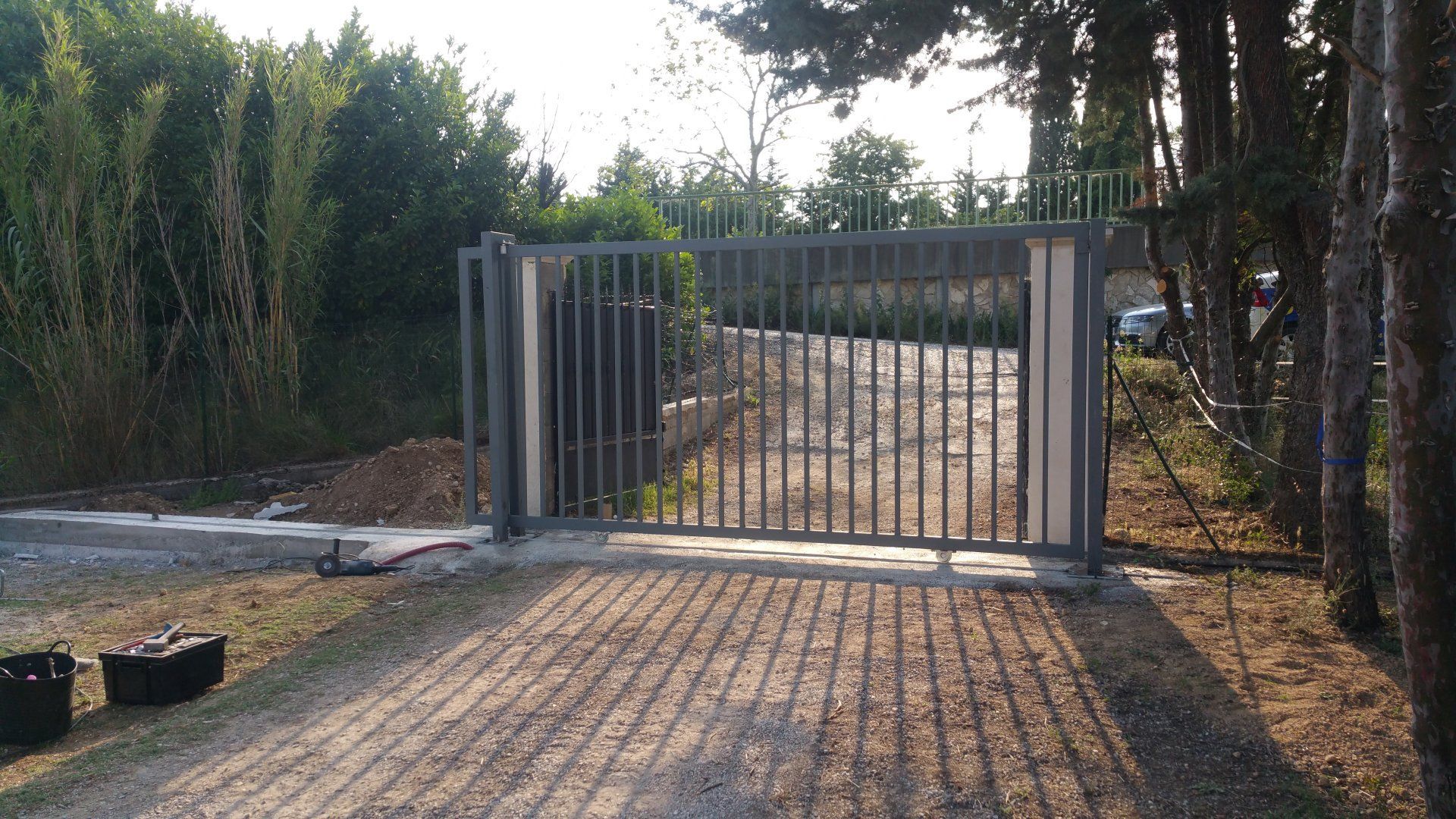 Un portail métallique bloque une allée de gravier, bordée d'arbres et de buissons baignés de soleil.
