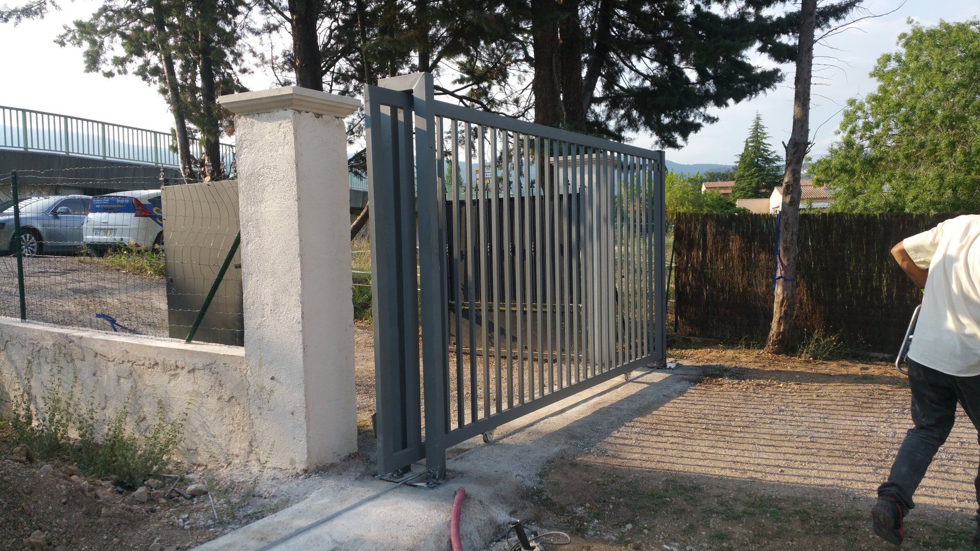 Portail métallique à côté d'un mur blanc et d'une allée, avec une personne partiellement visible à droite.