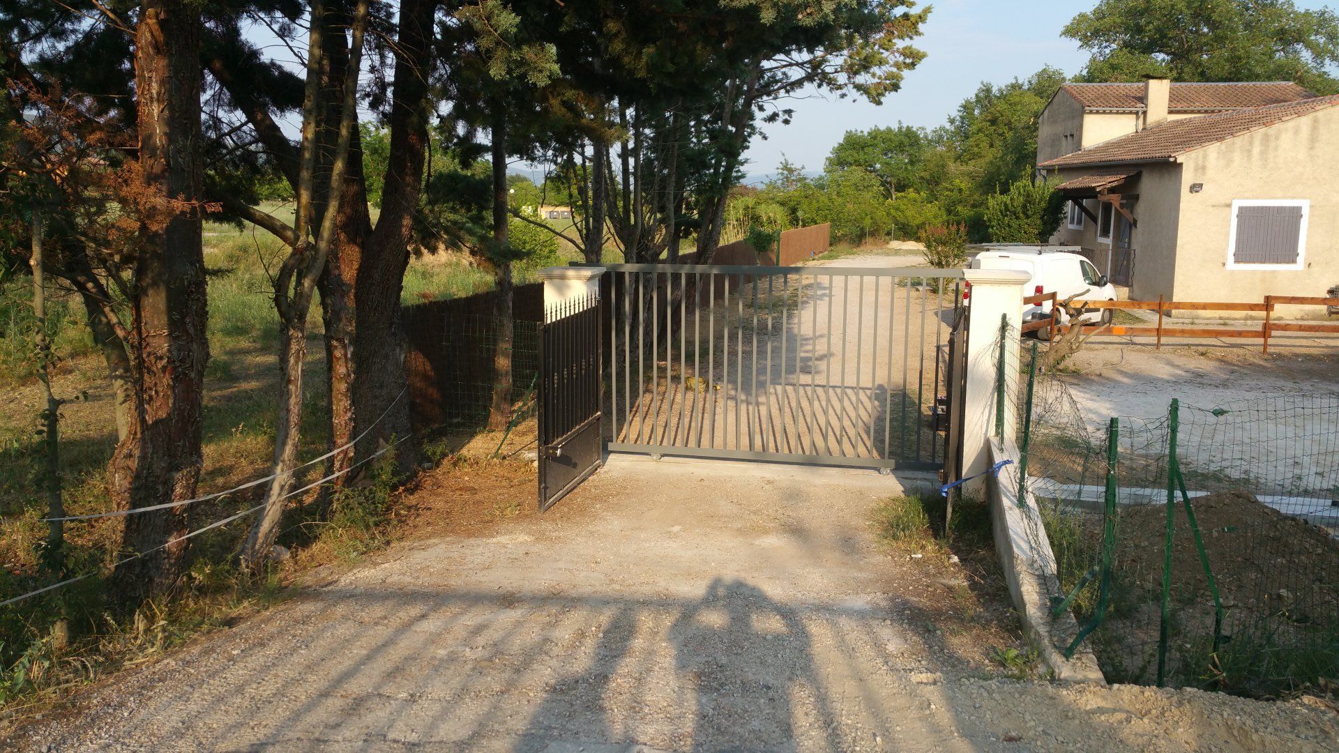 Allée de gravier menant à un portail métallique à côté d'une maison et d'arbres baignés d'un soleil chaud