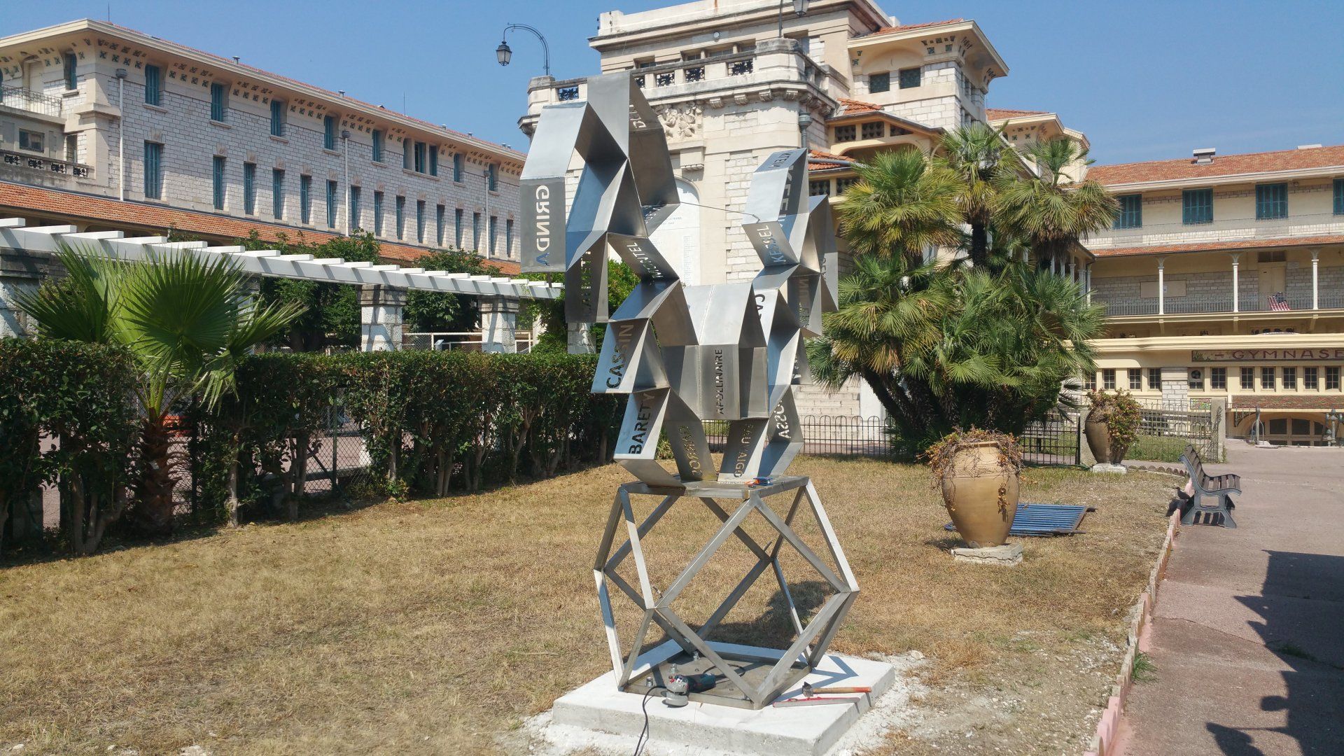 Sculpture géométrique en métal sur une pelouse entre des bâtiments de style campus, sous un ciel bleu clair.