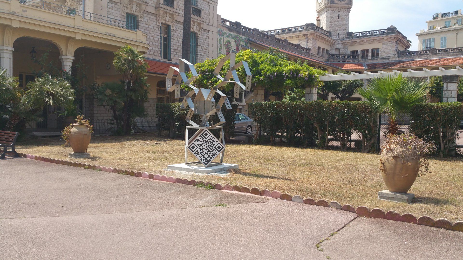Cour intérieure avec une statue sur un piédestal blanc, entourée de plantes en pot et de bâtiments en pierre sous un ciel ensoleillé