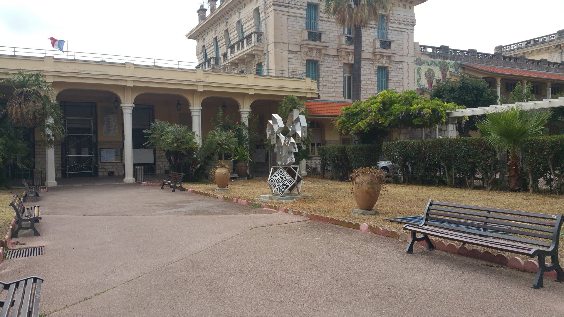 Cour intérieure avec bâtiment en pierre, allée couverte, sculpture abstraite, bancs et plantes en pot.