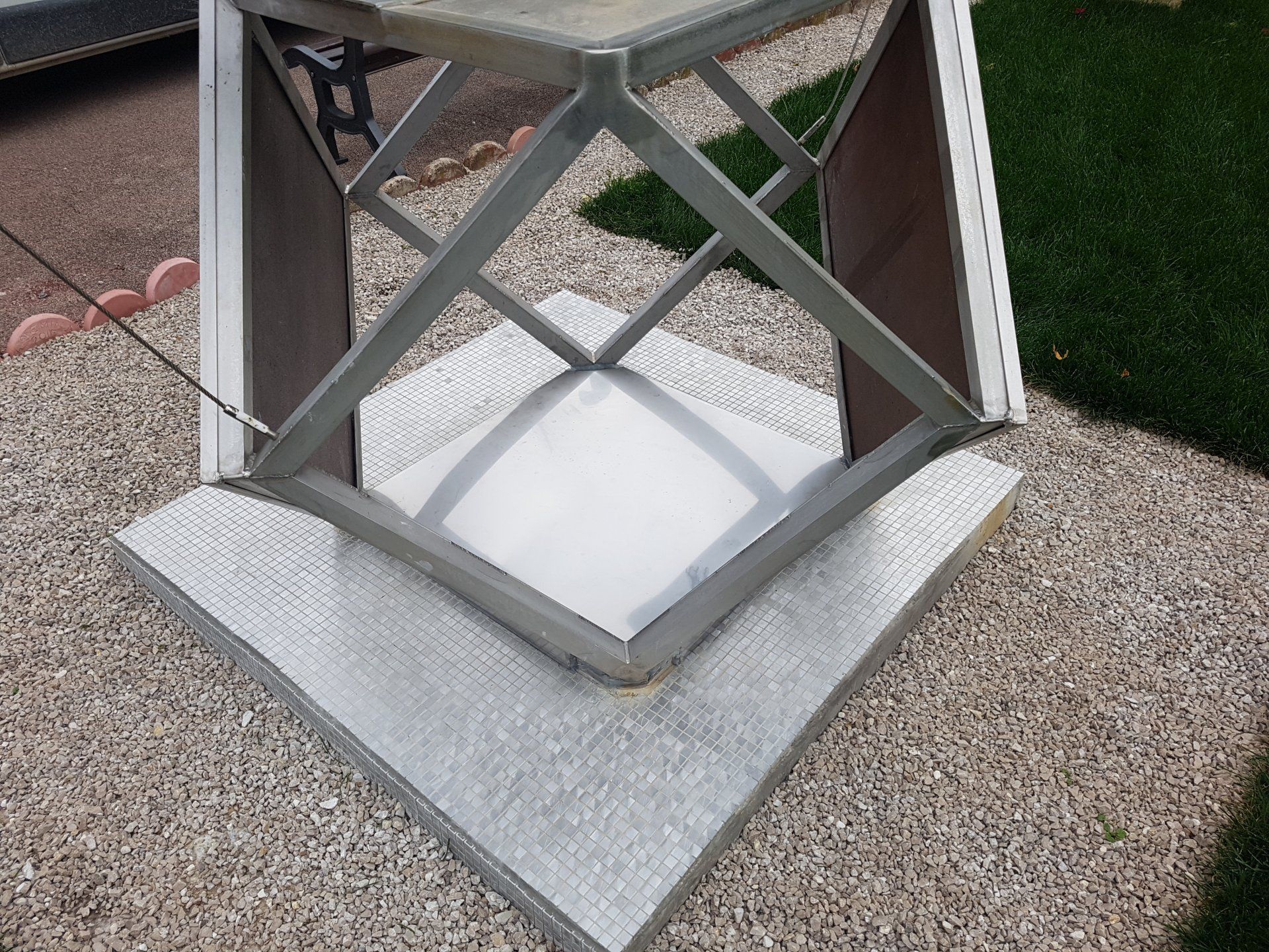 Socle en treillis métallique monté sur un piédestal en béton dans une cour gravillonnée.