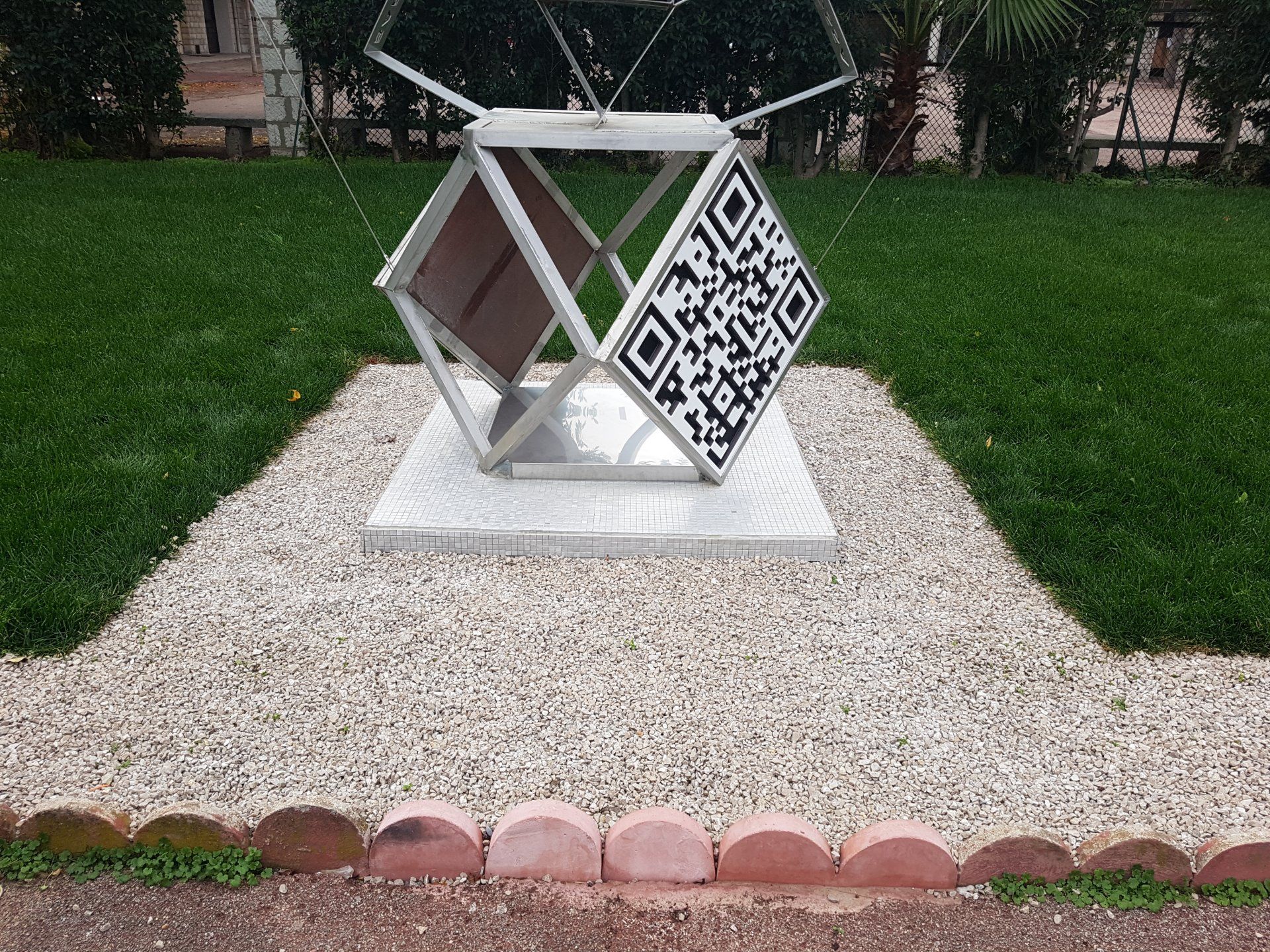 Sculpture décorative géométrique en métal sur un socle blanc, posée sur du gravier entourée d'herbe verte.