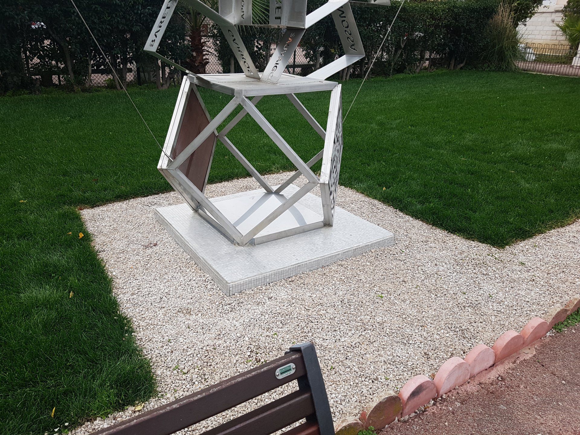 Sculpture géométrique en métal sur une dalle de béton dans un jardin paysager avec allée de gazon et de gravier
