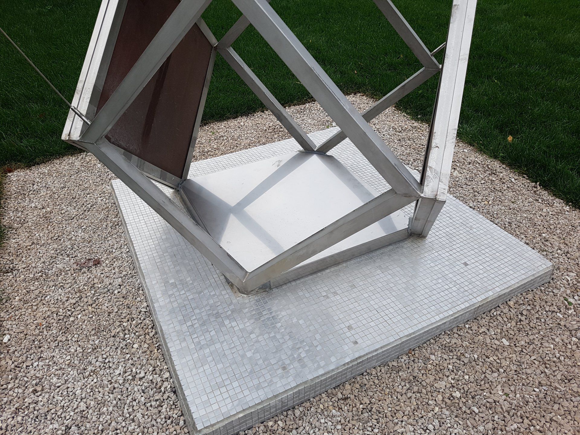 Une sculpture géométrique blanche en extérieur, sur un socle en béton, à côté de gravier et d'herbe.