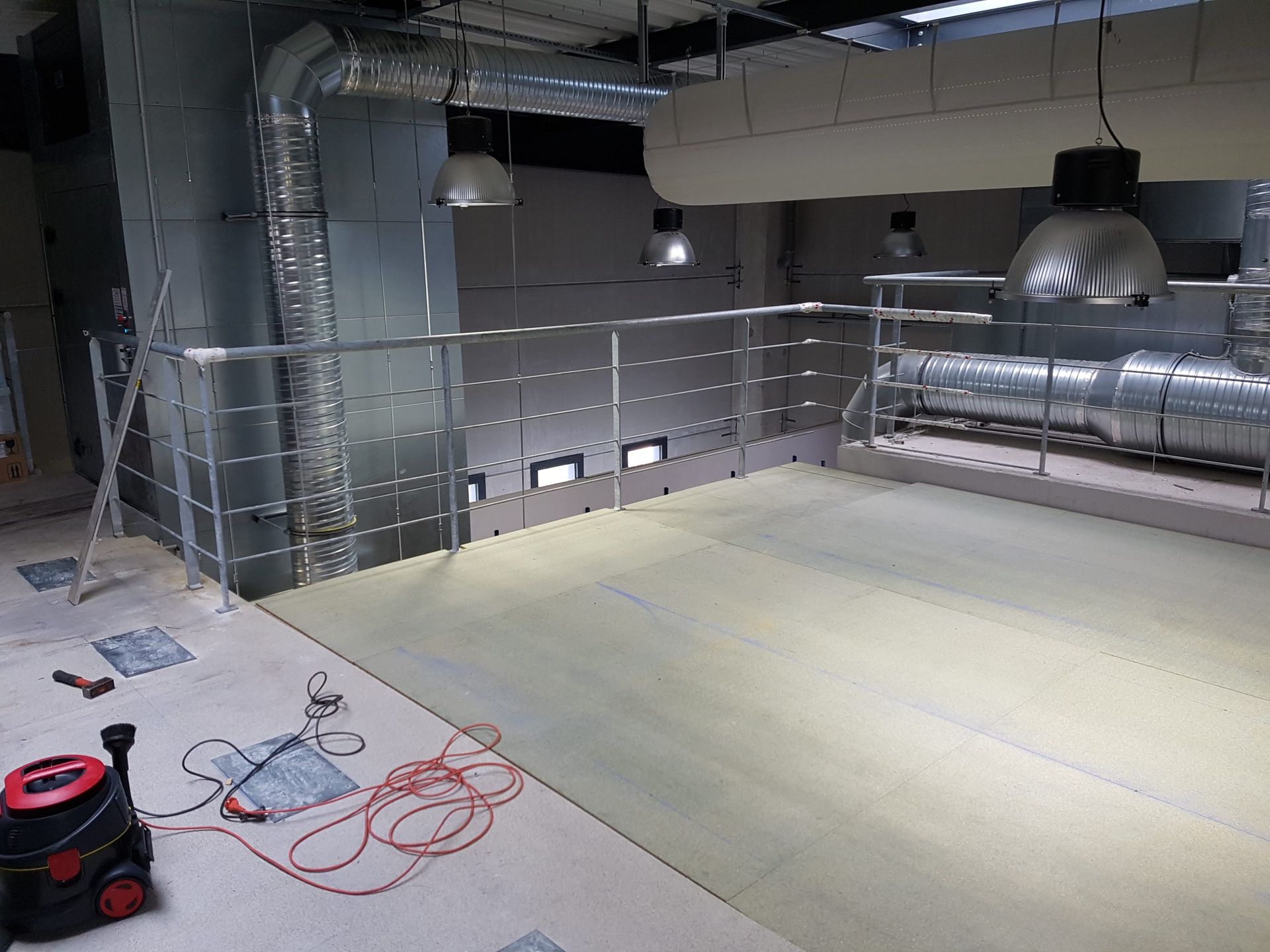 Pièce industrielle vide avec conduits apparents, luminaires suspendus et câbles orange enroulés au sol