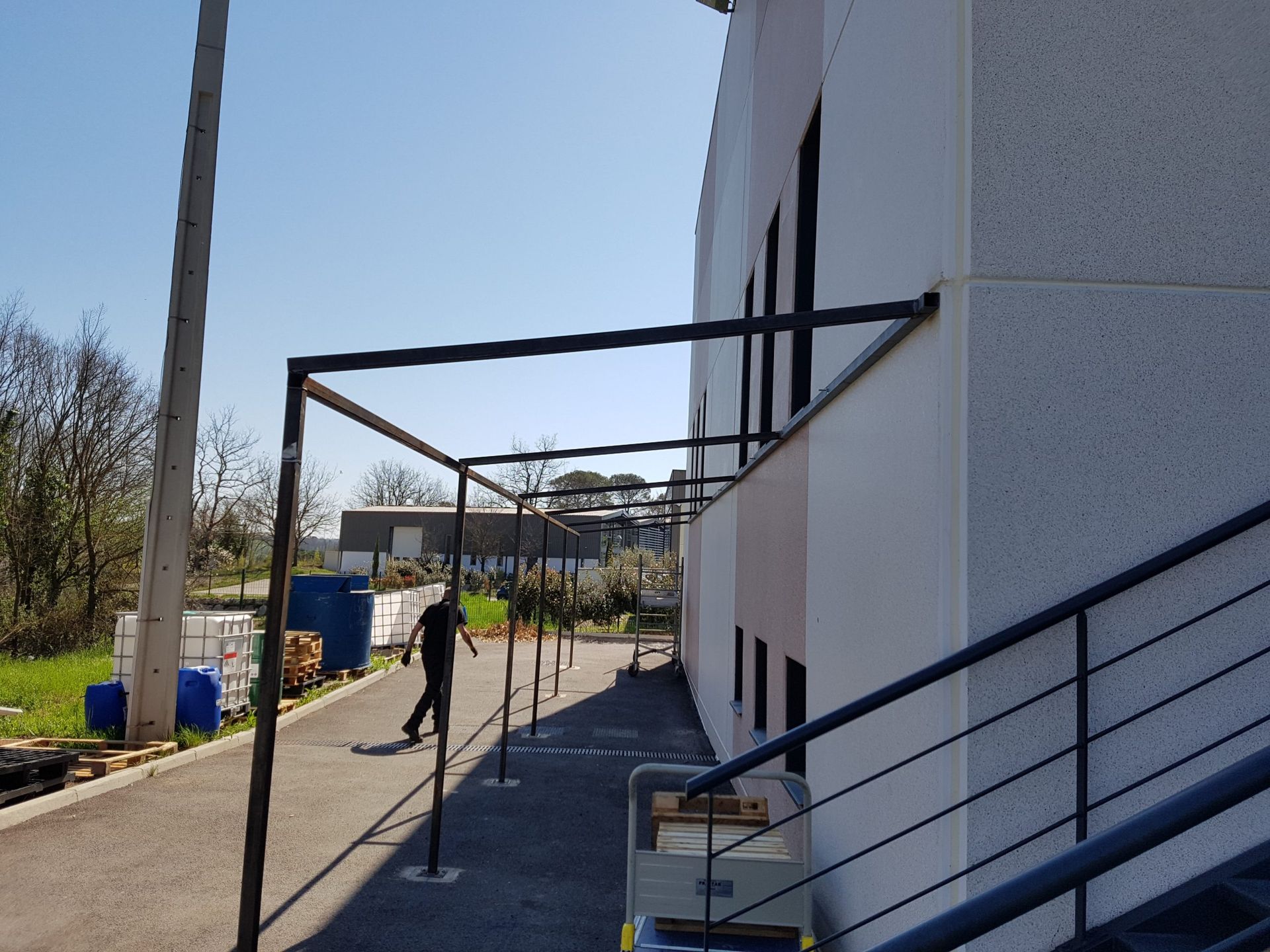 Allée extérieure longeant un bâtiment blanc, avec des rambardes métalliques, des poteaux et quelques personnes au loin.