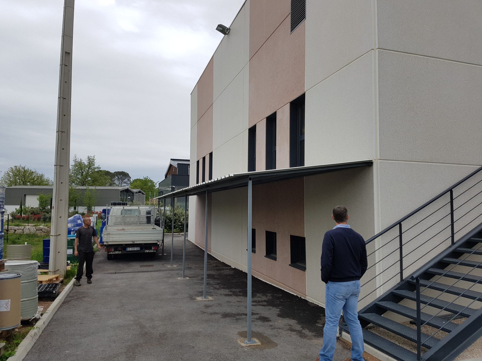 Des gens marchent à côté d'un bâtiment beige avec des escaliers et un camion garé sous un ciel couvert.