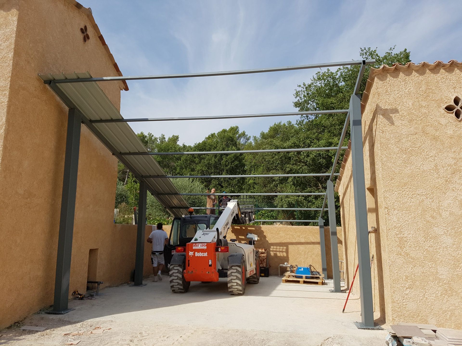 Des engins de chantier sous un auvent métallique entre des murs en stuc dans une cour