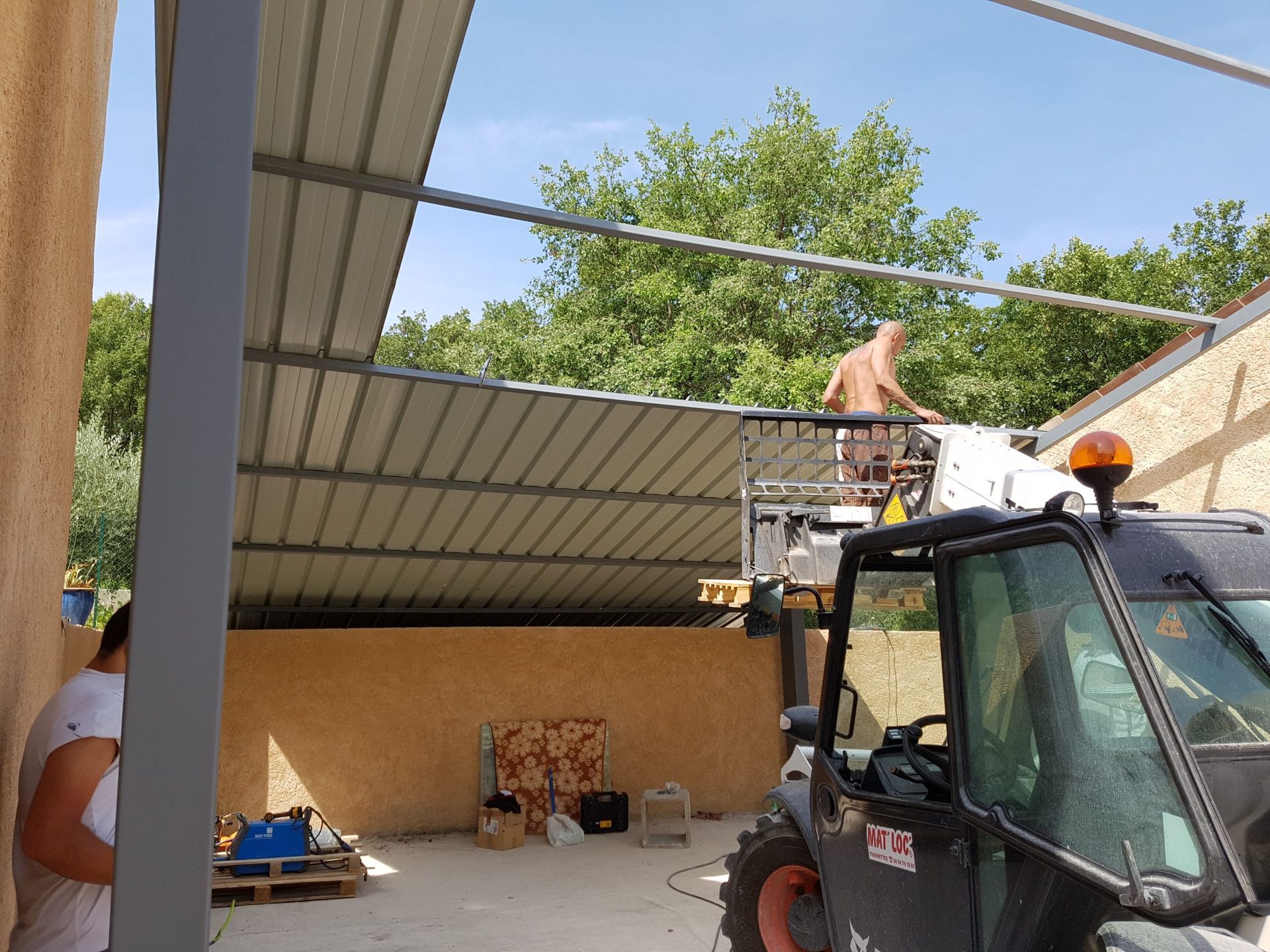 Des ouvriers installent une toiture métallique au-dessus d'une terrasse jouxtant une maison, tandis que des échafaudages et des matériaux de construction se trouvent à proximité.