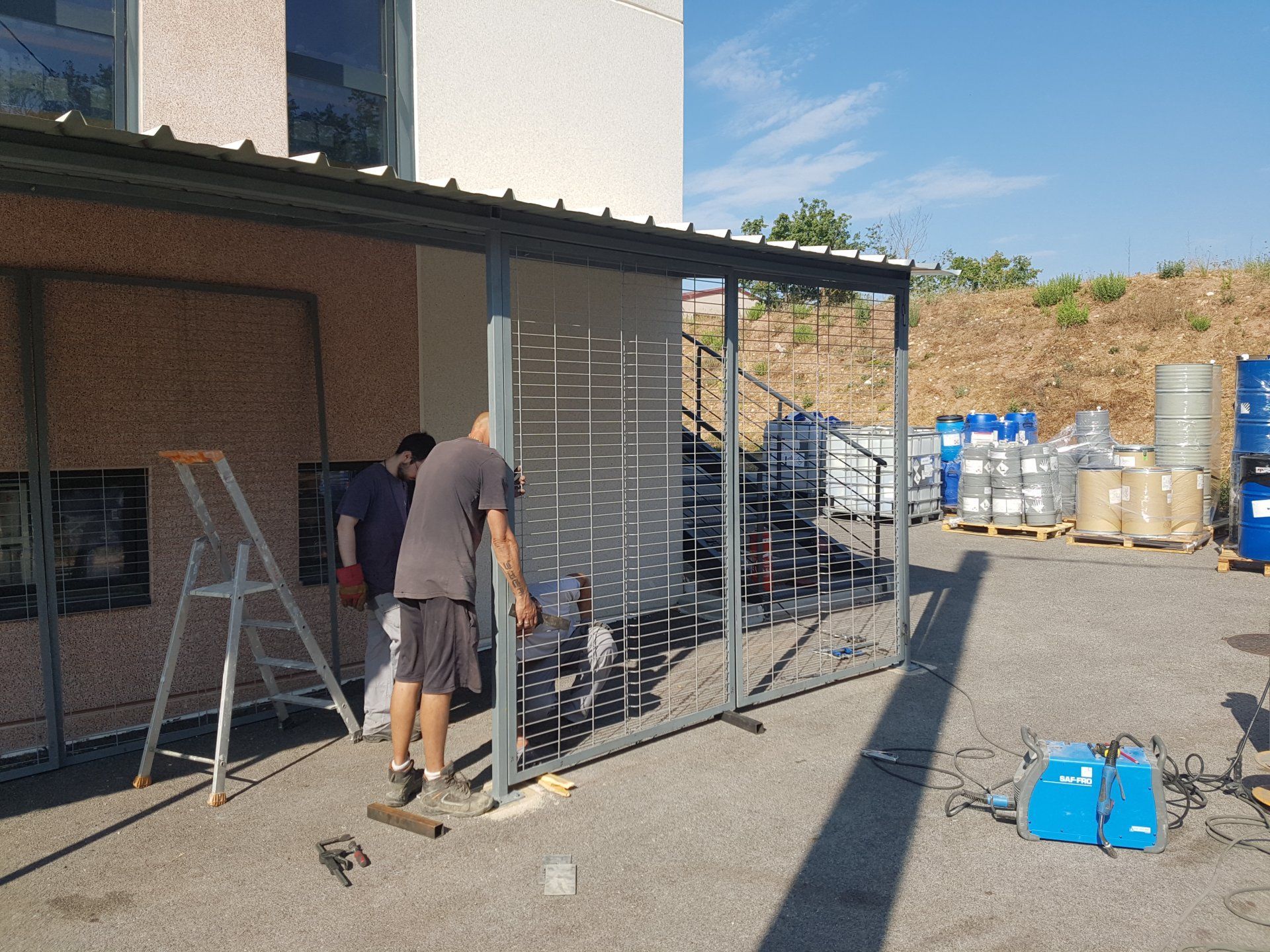 Deux personnes travaillent près d'une clôture métallique à l'extérieur d'un bâtiment, avec des échelles et du matériel empilé à proximité.