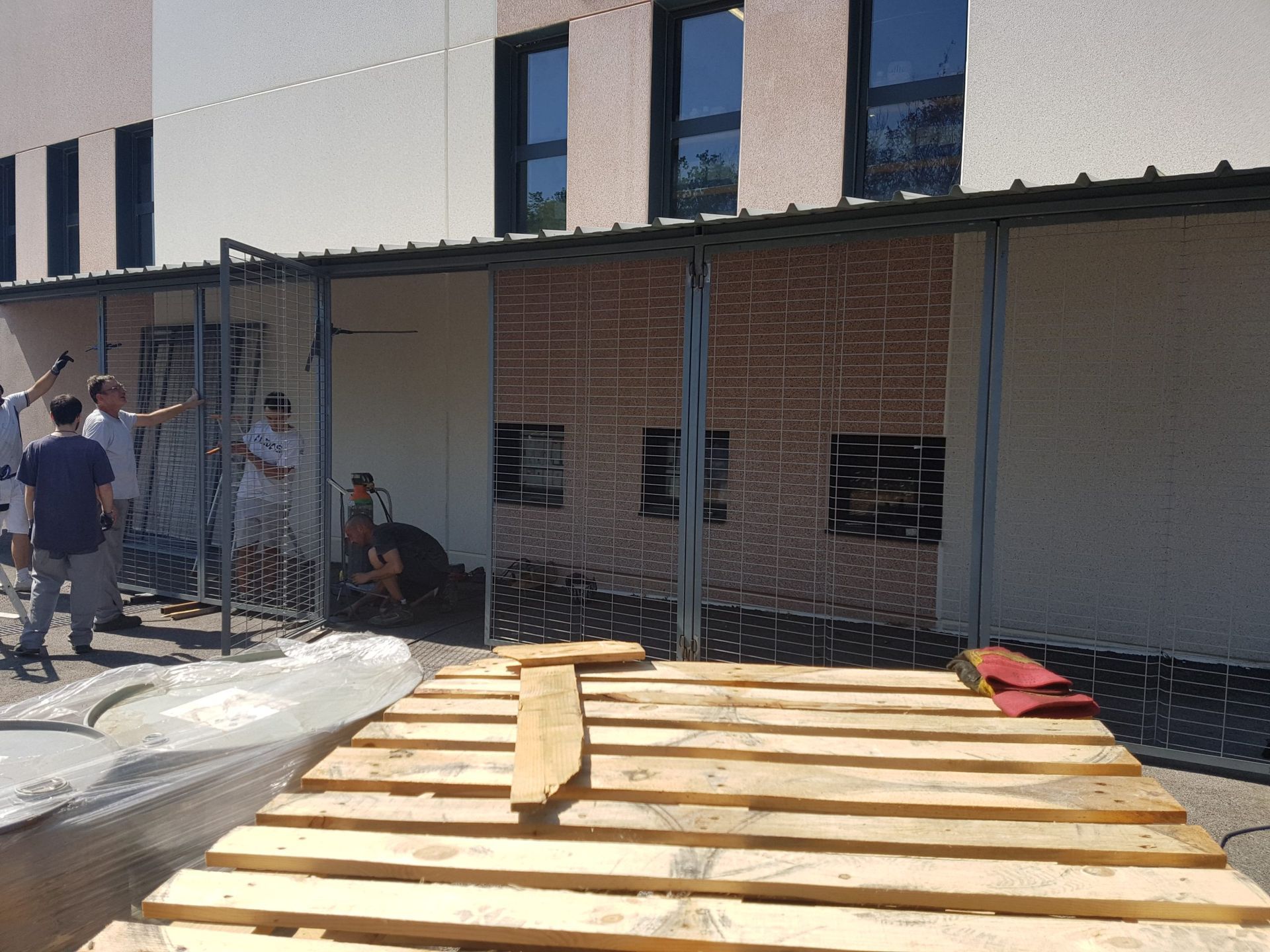 Zone de construction d'une terrasse extérieure avec clôture, ouvriers et palette en bois devant un bâtiment en briques.