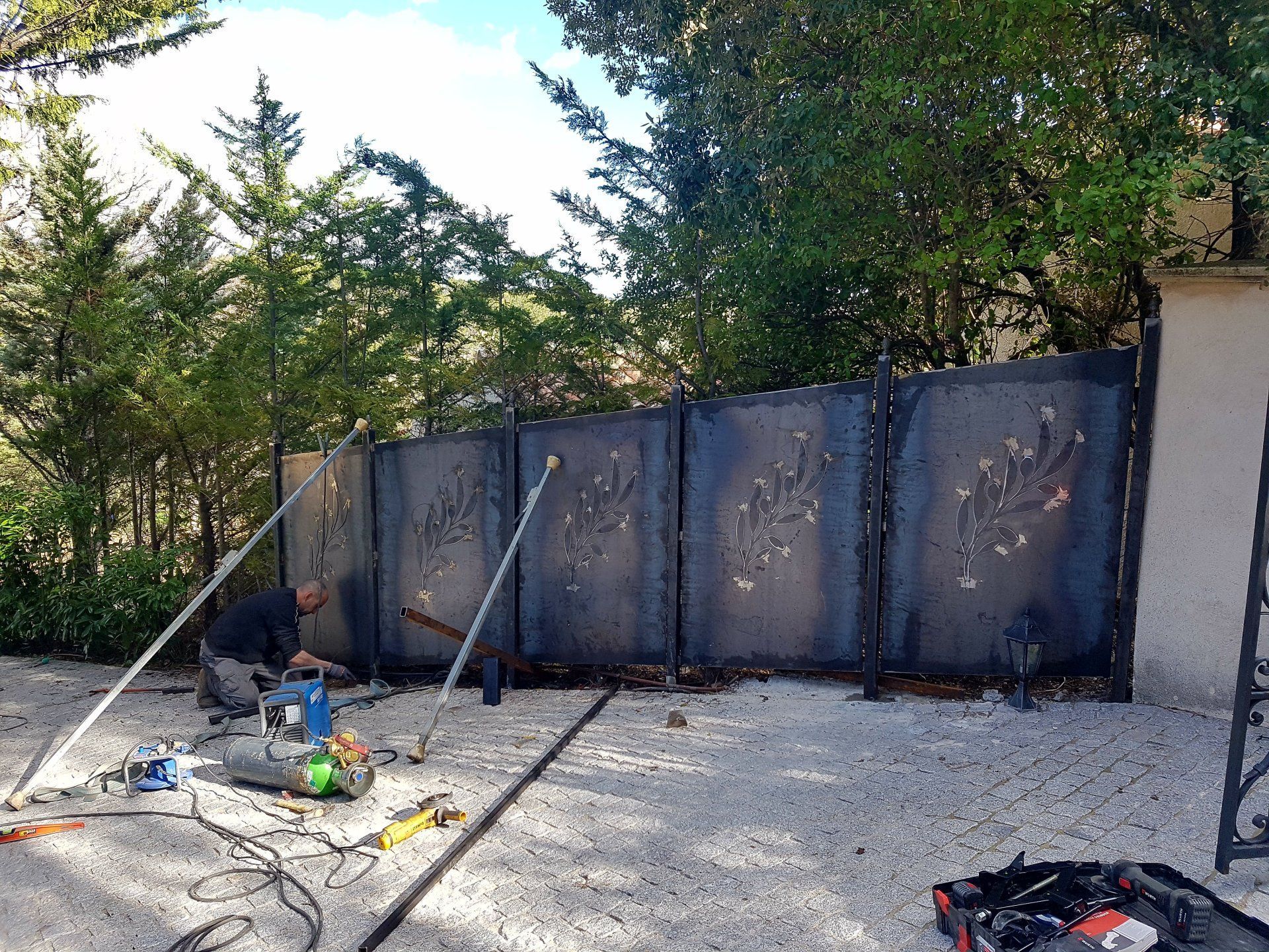 Zone de travail extérieure avec panneaux de clôture gris, arbres et outils de construction éparpillés sur du gravier.
