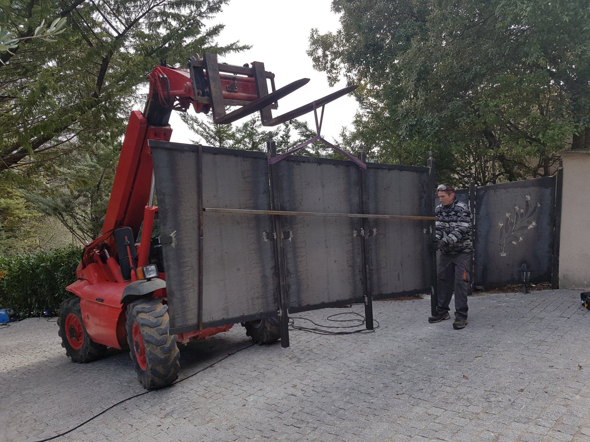 Un chariot élévateur rouge soulève un grand portail métallique à l'extérieur, un ouvrier se tenant à côté.