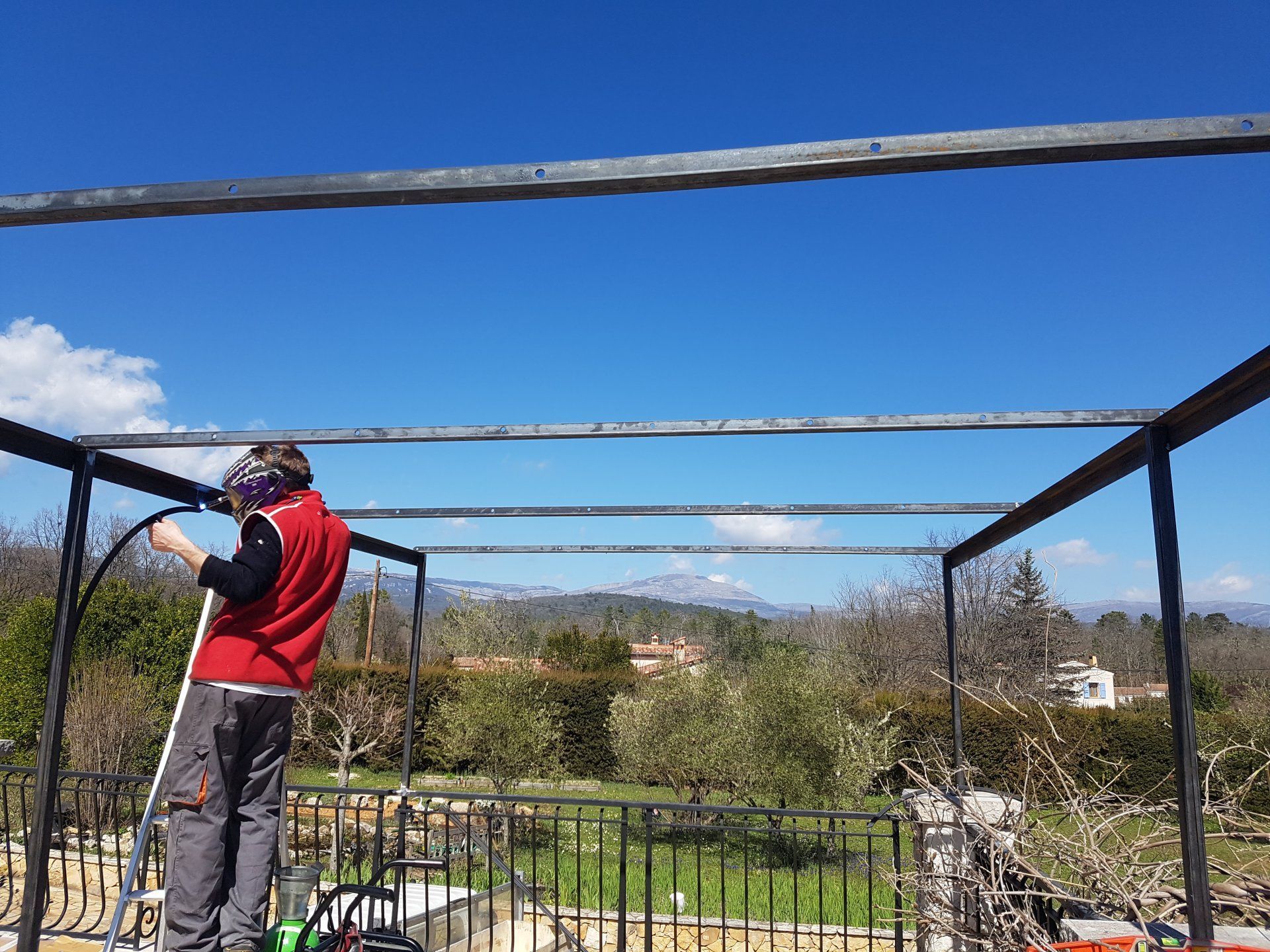 Une personne en veste rouge se tient sur une terrasse, regardant à travers une balustrade un paysage ensoleillé.
