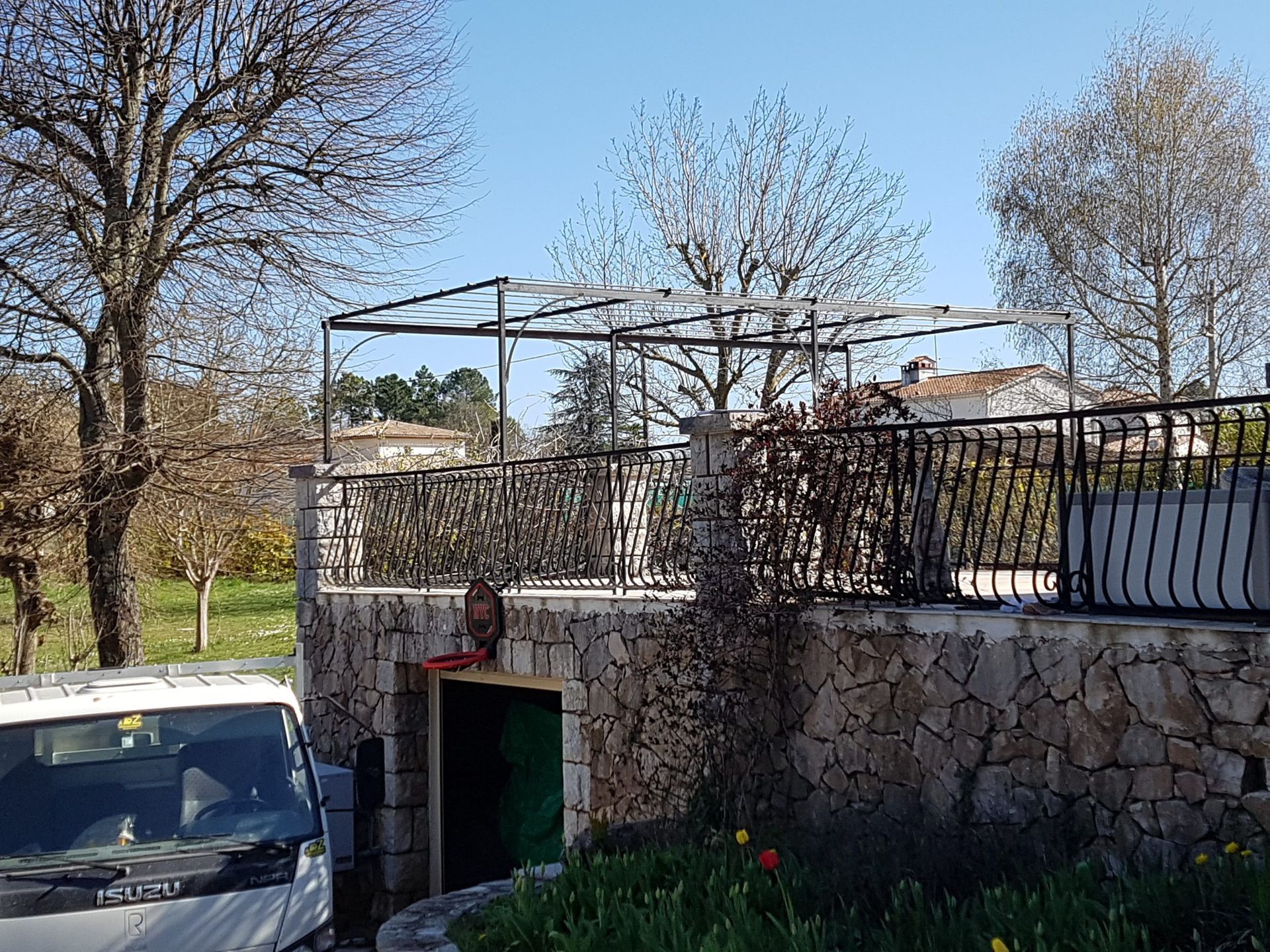 Mur en pierre avec une porte verte et une terrasse sur le toit, à côté d'une voiture garée sous des arbres dénudés.