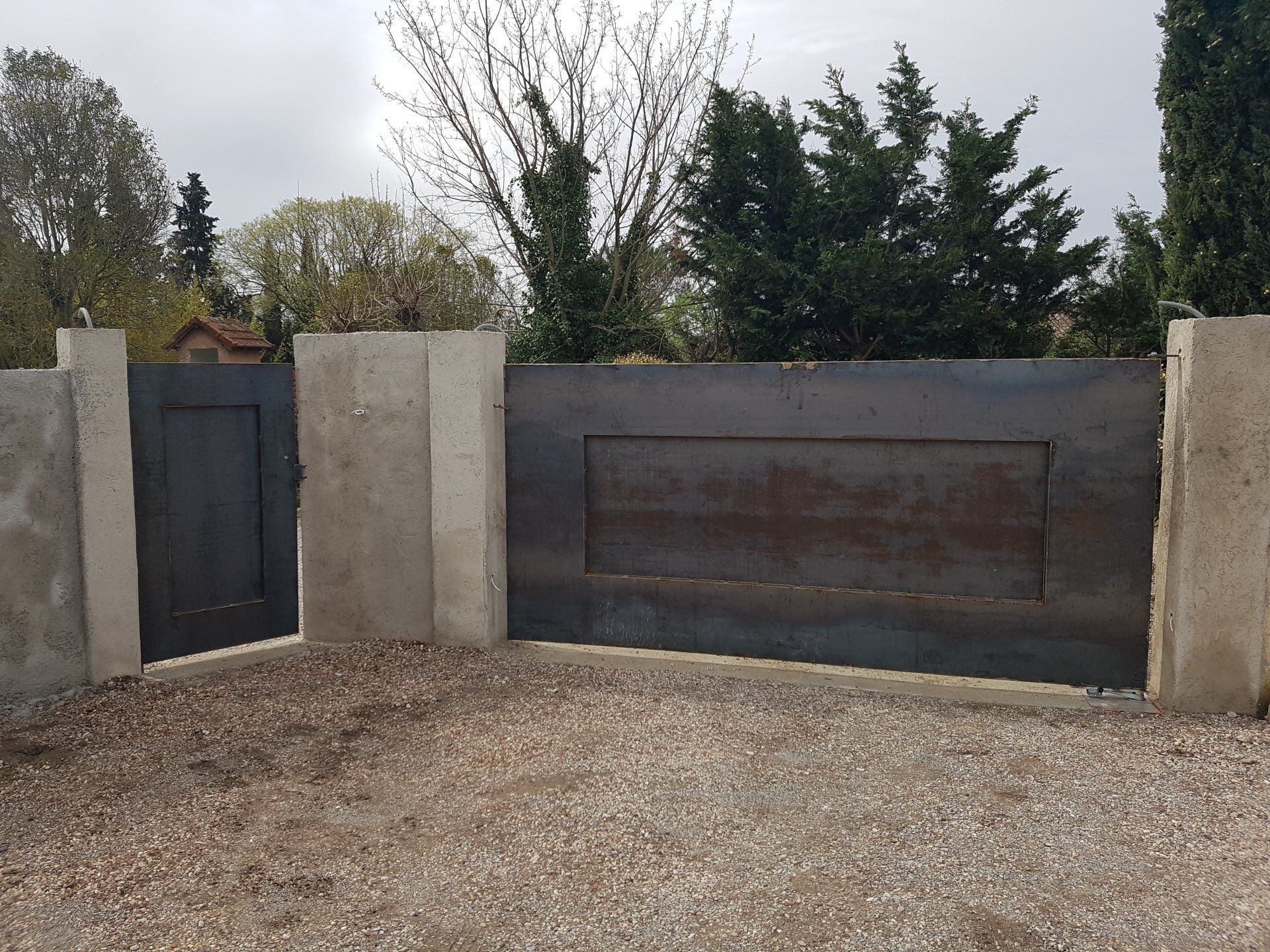 Mur en béton avec un portail noir et un grand panneau métallique bordant une allée de gravier sous les arbres