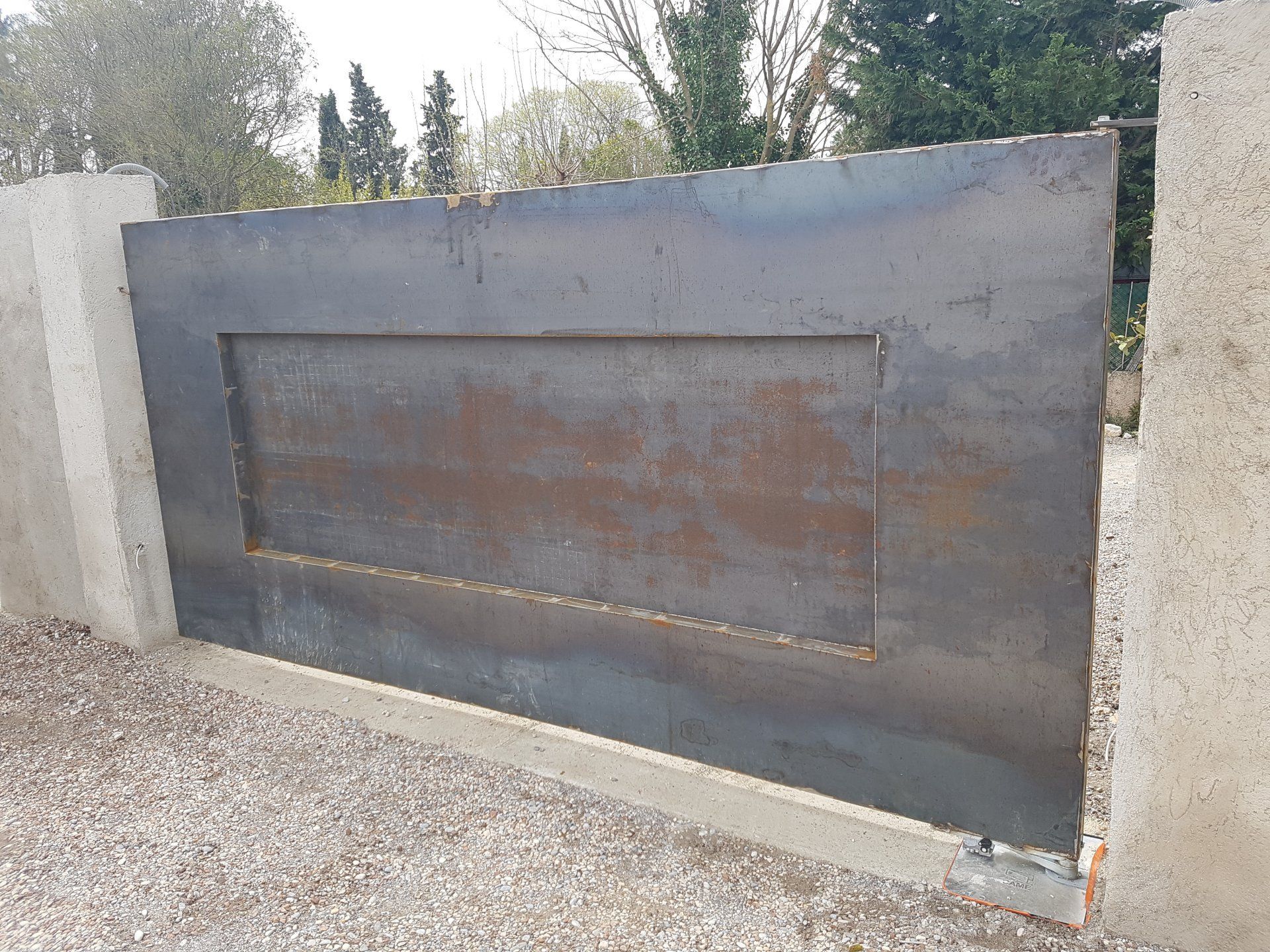 Grand portail en métal noir avec un panneau rectangulaire rouillé, installé dans une allée de gravier.