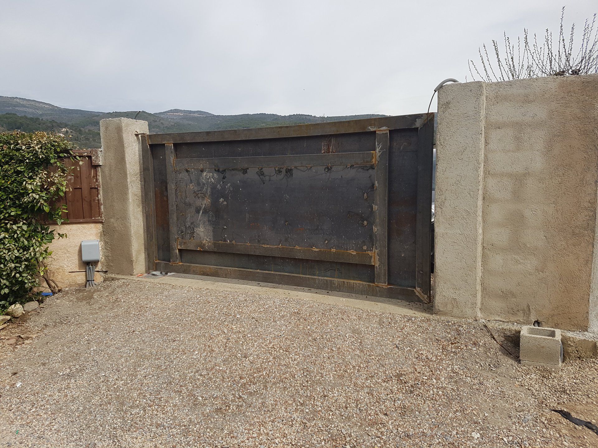 Portail en métal rouillé installé dans une allée de gravier entre des murs en béton, avec une colline en arrière-plan.