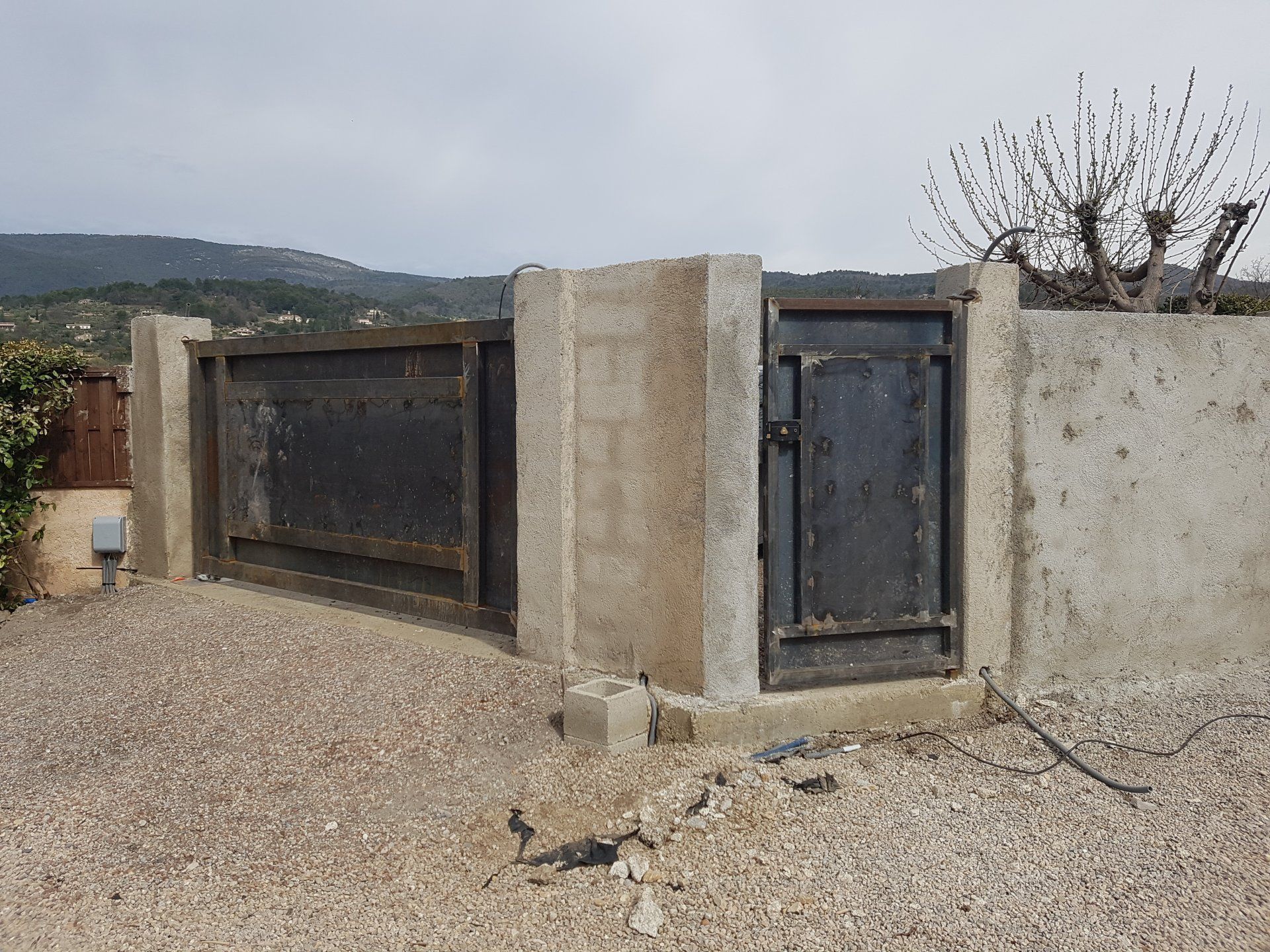 Mur en béton avec portails en métal noir sur une allée de gravier dans un cadre rural