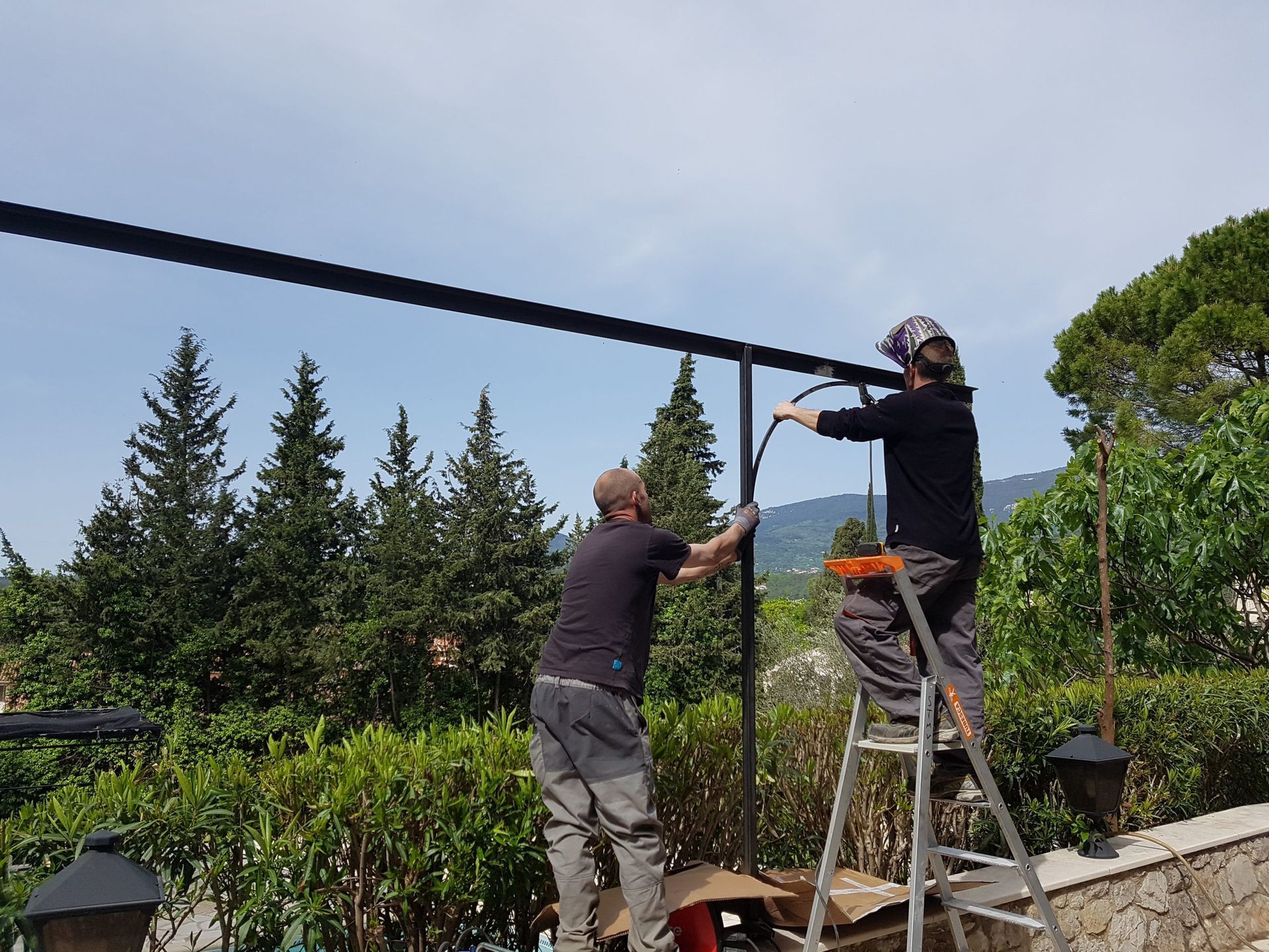 Deux ouvriers sur des échelles installent une structure métallique noire au-dessus d'une haie de jardin.