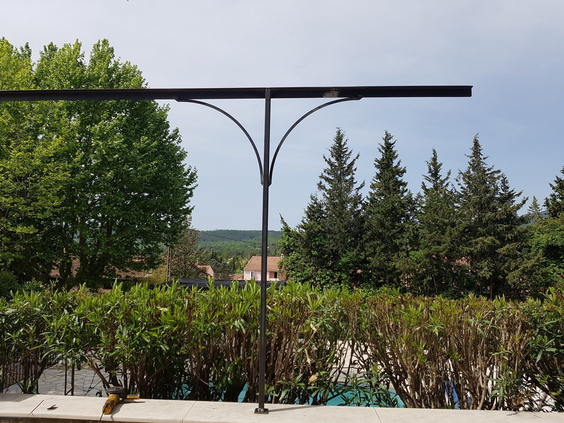 Parc avec haies, grands arbres et une pergola en métal sombre se détachant sur un ciel lumineux.