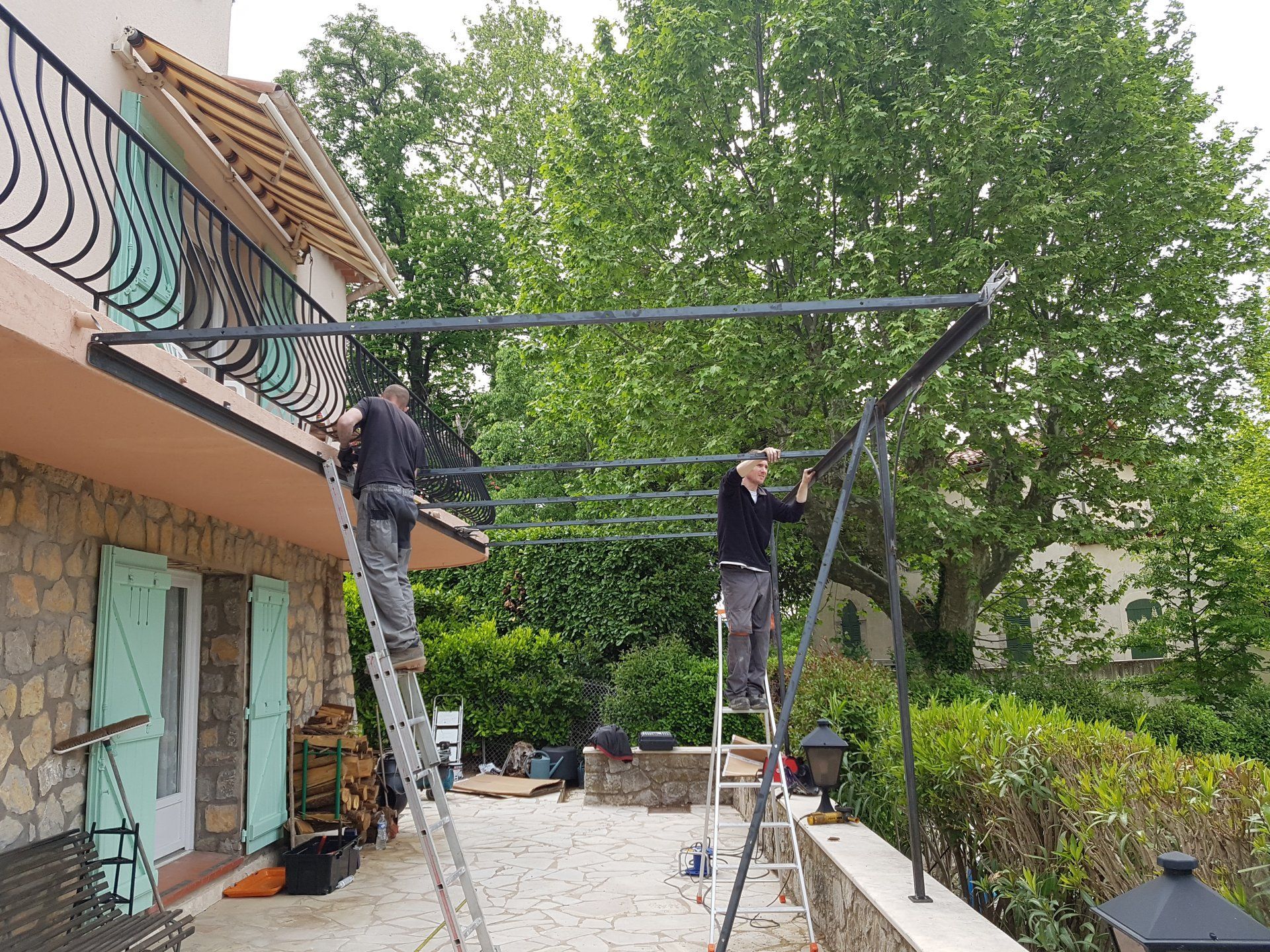 Deux ouvriers assemblent une structure métallique à l'extérieur, à côté d'une maison et d'arbres.