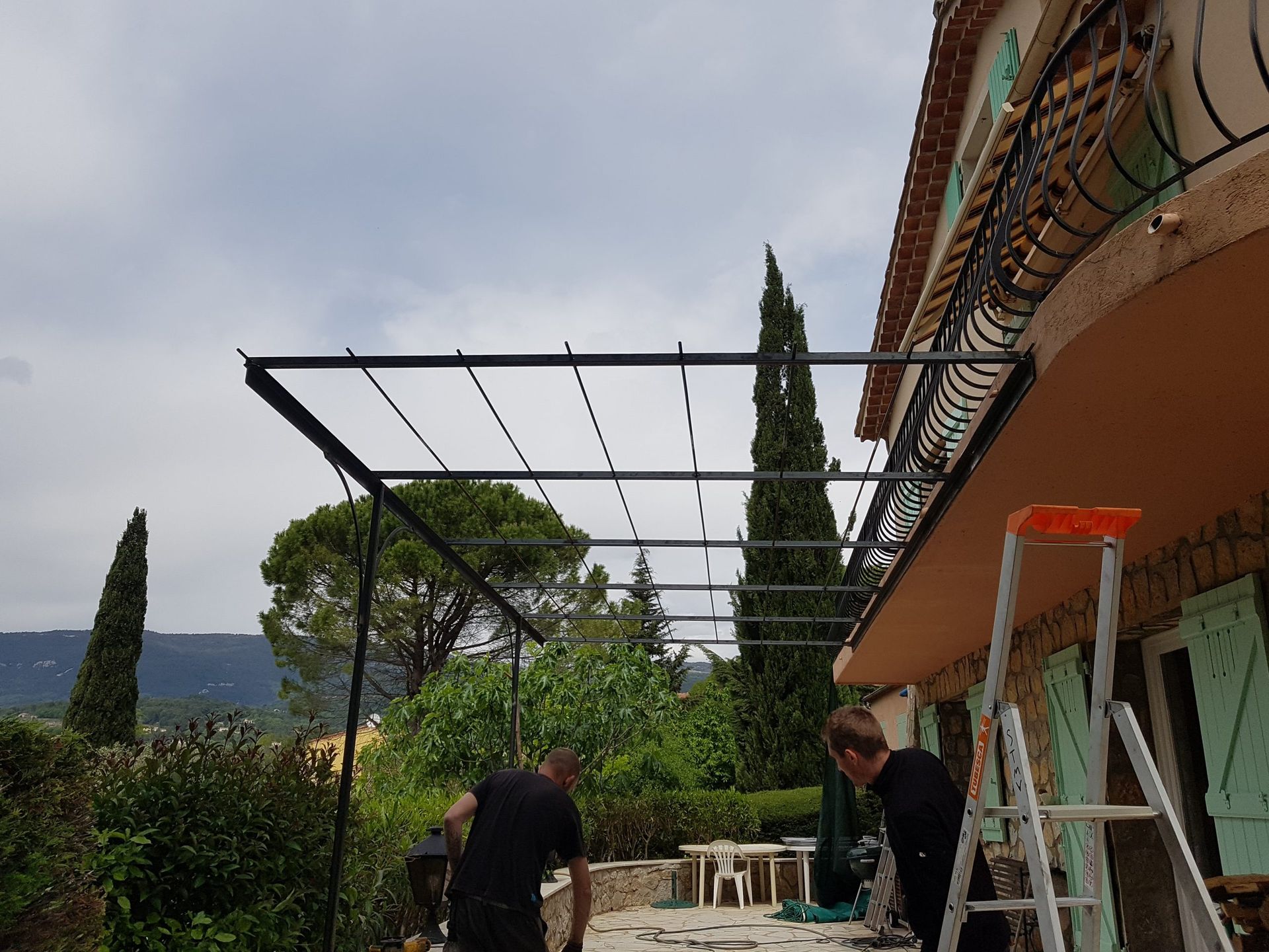 Des ouvriers travaillent sur la charpente d'un toit de terrasse à côté d'une échelle, avec des arbres et un ciel nuageux en arrière-plan.