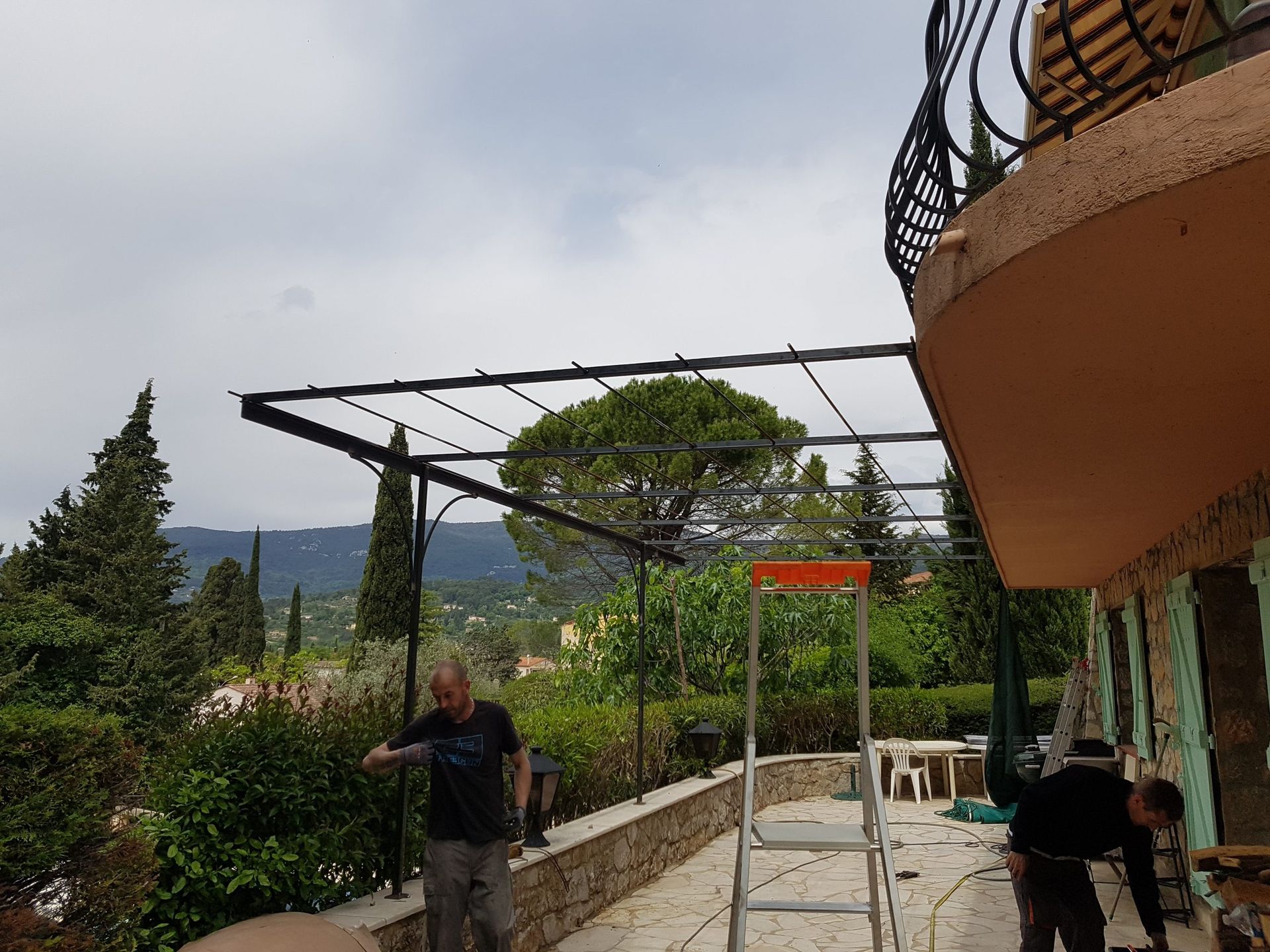 Une personne installe une pergola noire sur une terrasse avec de la verdure et des montagnes en arrière-plan.