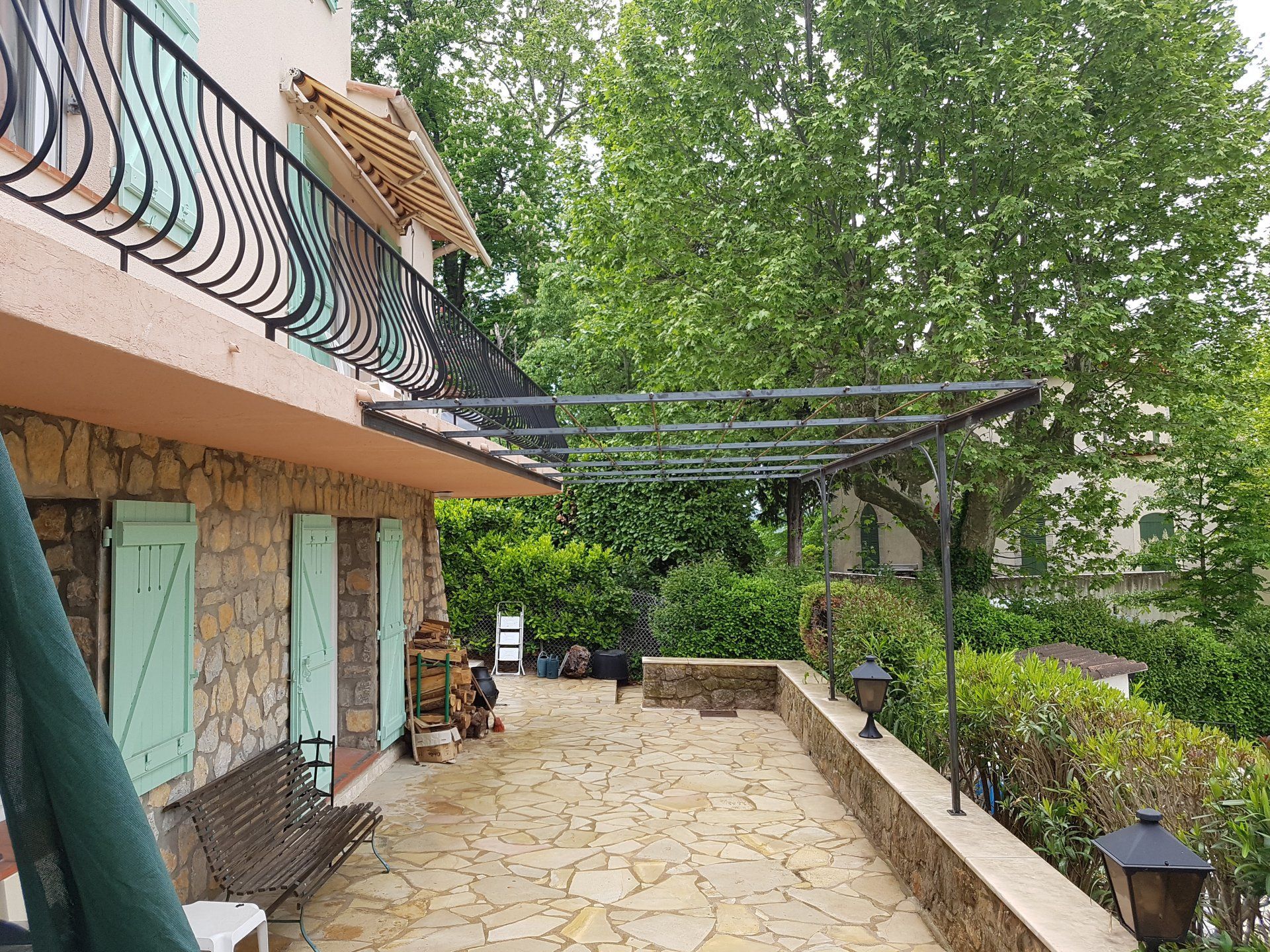 Terrasse en pierre jouxtant une maison avec des arbres verts, une pergola noire et une balustrade de balcon.