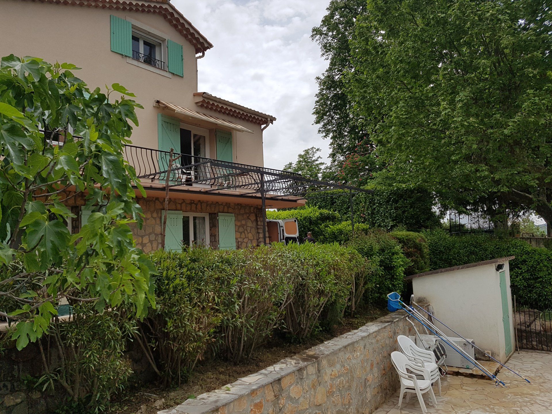 Maison de style méditerranéen avec volets verts, haie de jardin et chaises de patio au bord d'une allée bordée d'arbres.