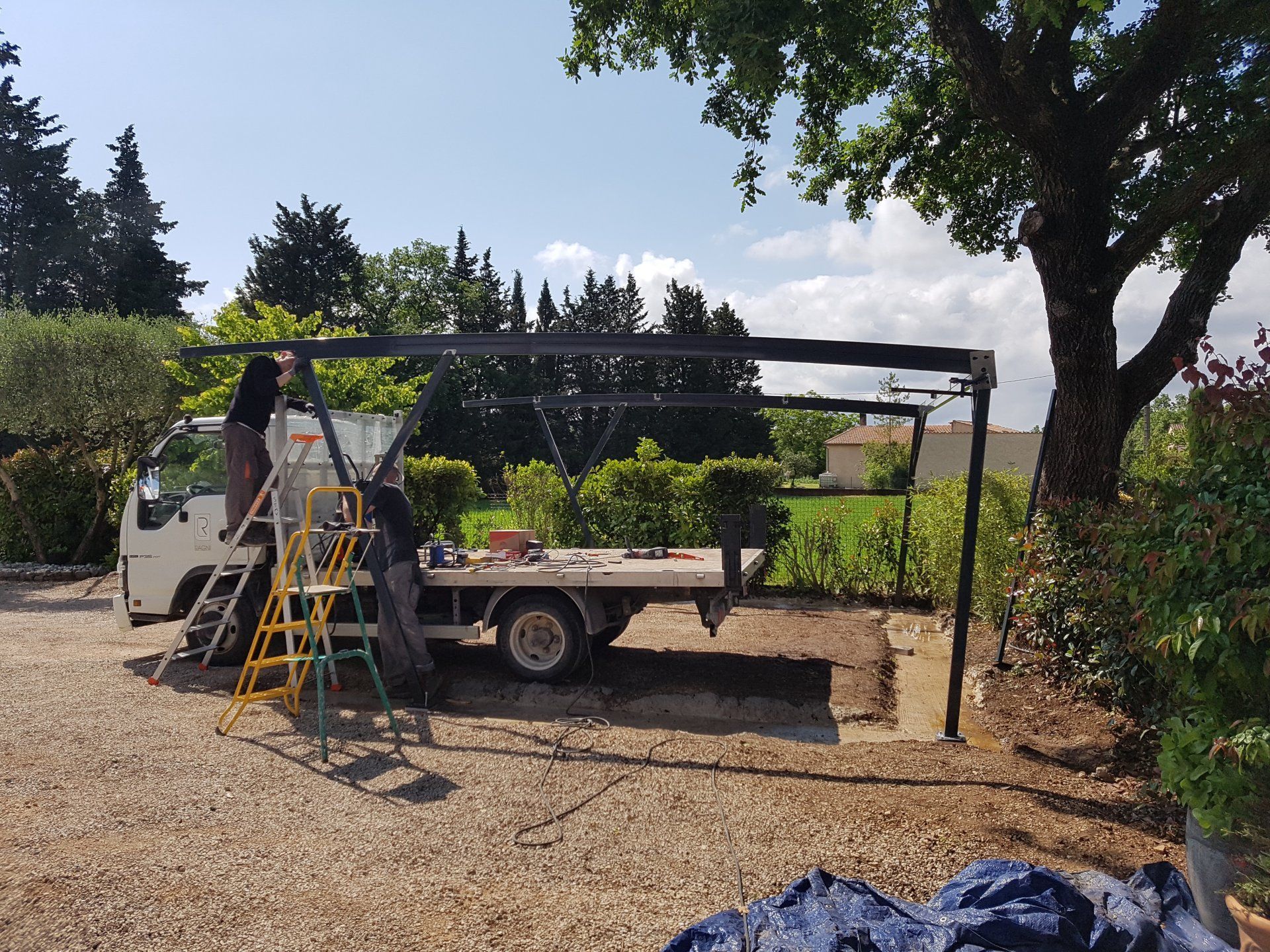 Un ouvrier sur une échelle à côté d'un camion sous un auvent, dans une cour gravillonnée avec des plantes et des arbres.