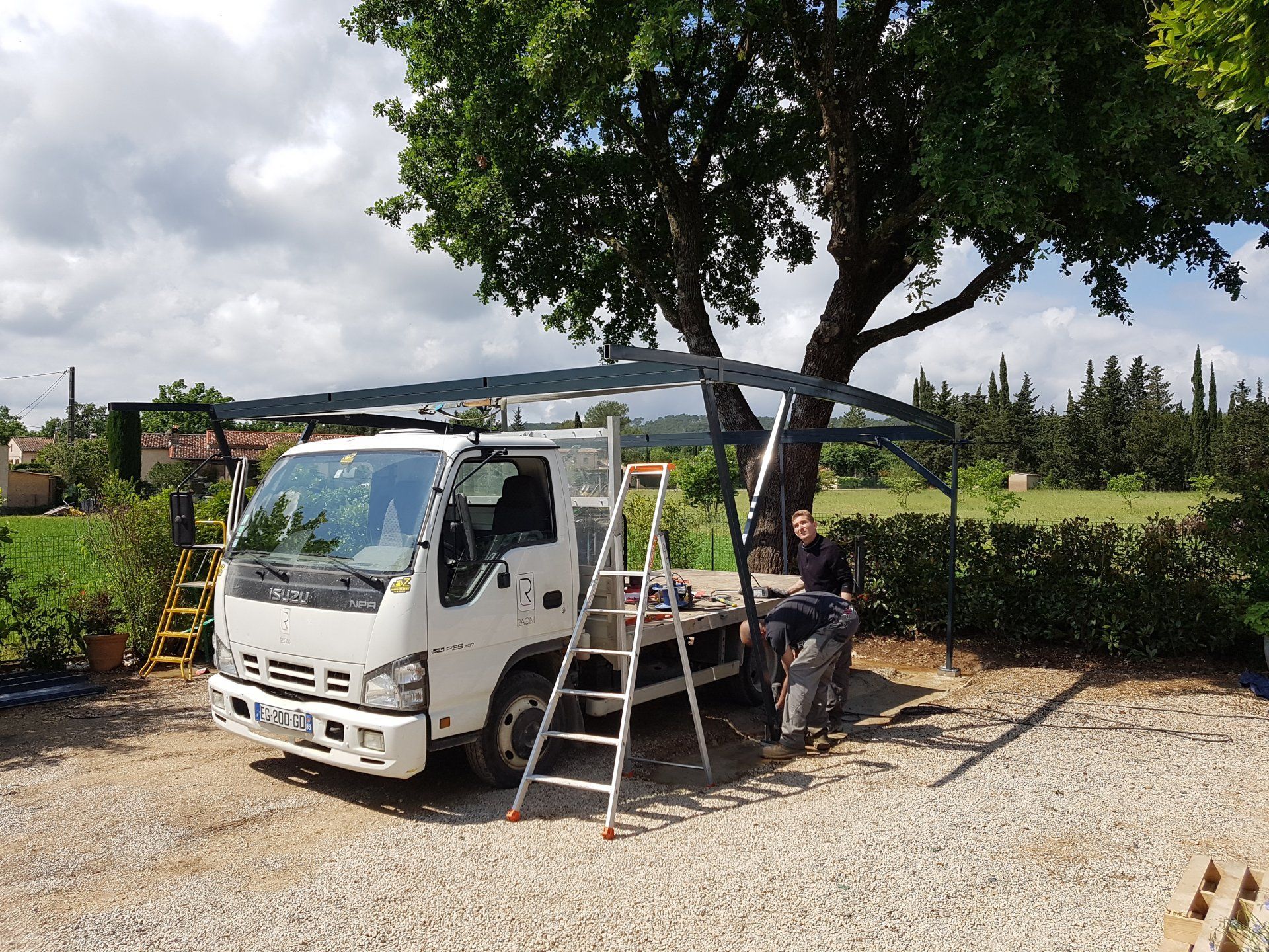 Un ouvrier sur une échelle à côté d'un camion utilitaire blanc sous un grand arbre, sur un terrain gravillonné.