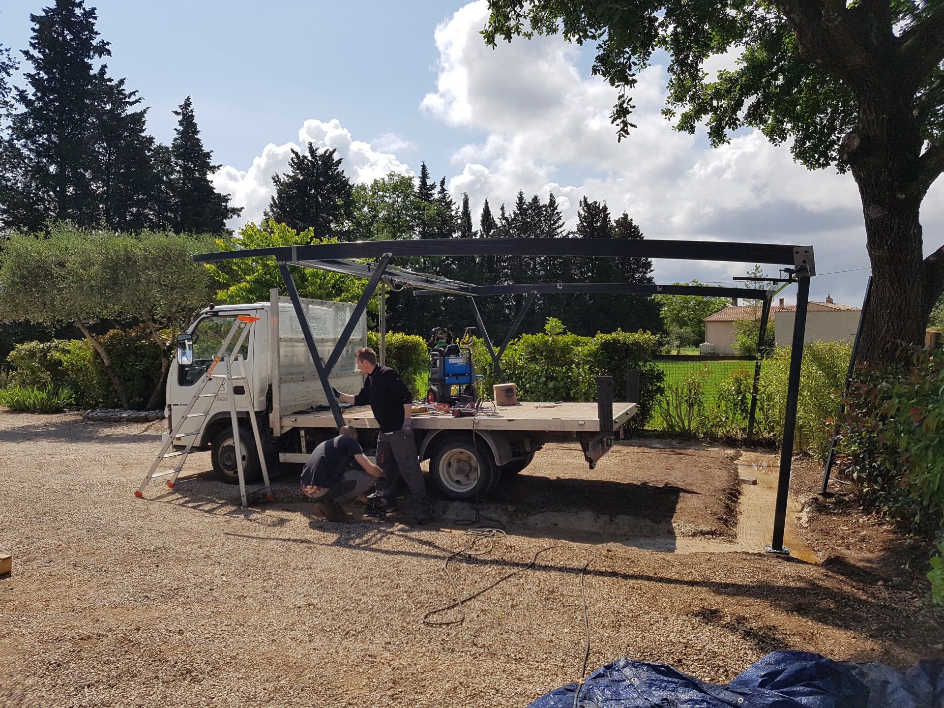 Une personne travaille à côté d'un petit camion utilitaire blanc sous un abri dans une cour gravillonnée.