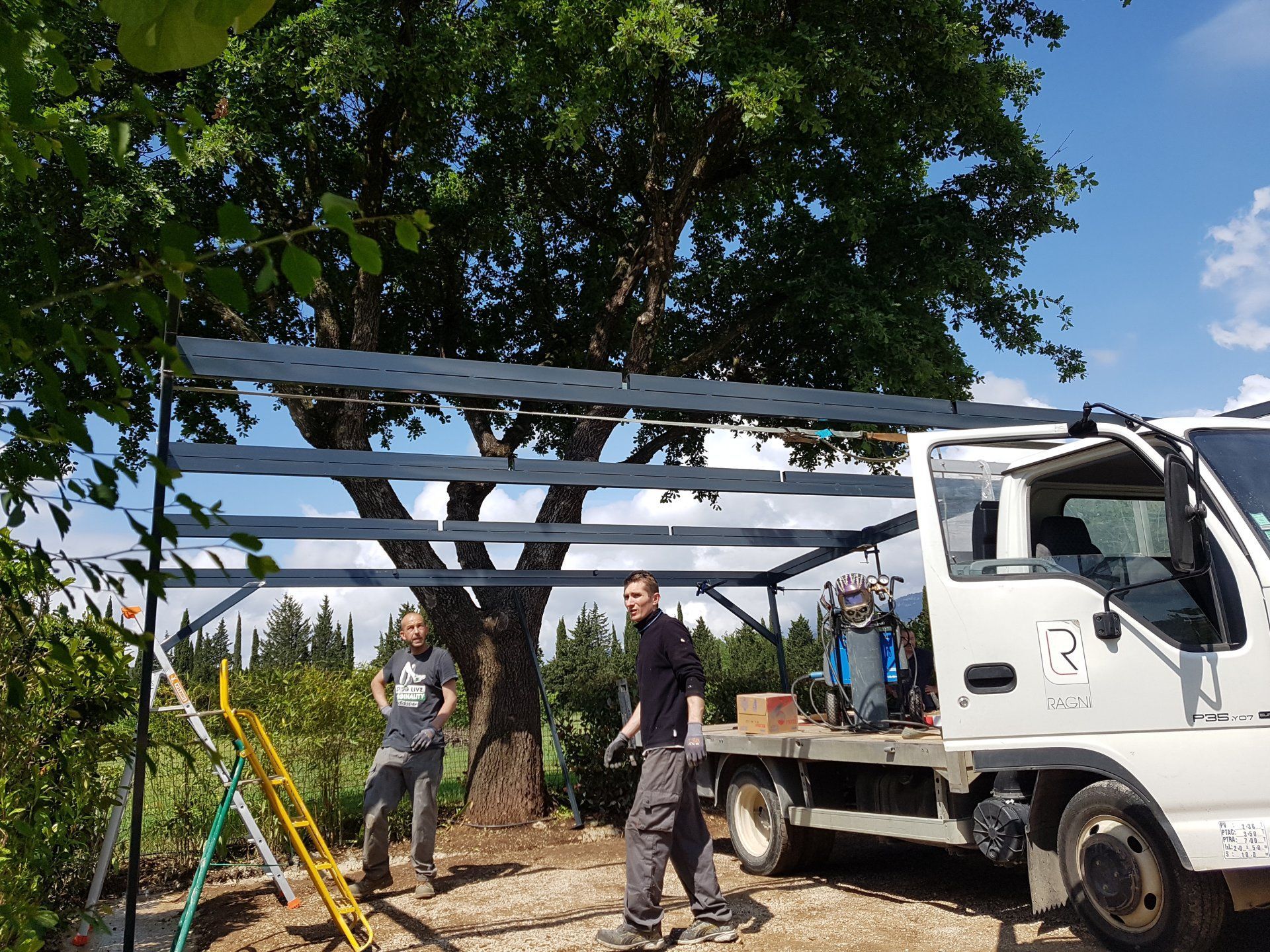 Des ouvriers assemblent une structure métallique à côté d'un camion blanc, sous un grand arbre, par une journée ensoleillée.