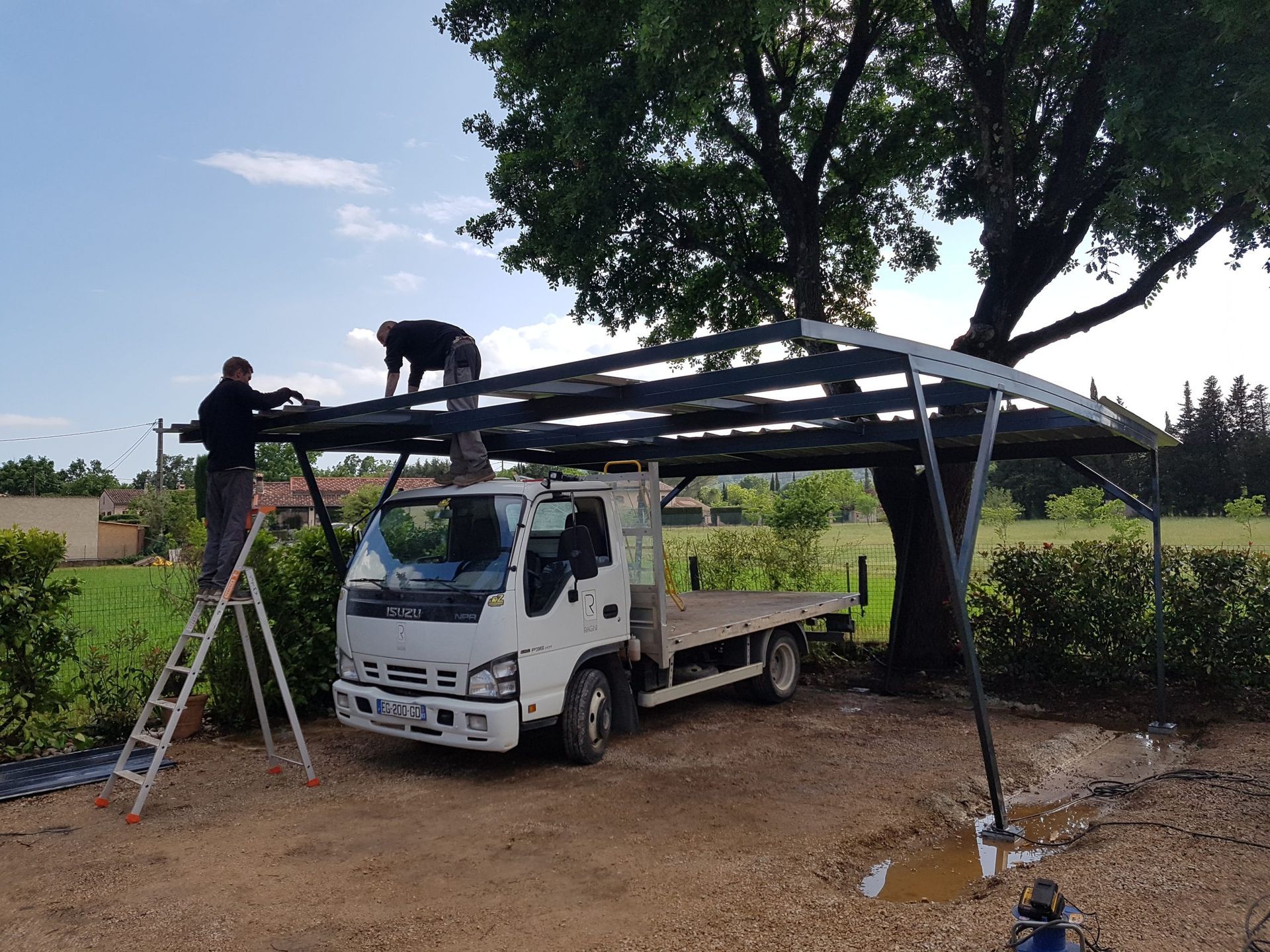 Deux ouvriers installent une charpente métallique sur un camion blanc sous un grand arbre.