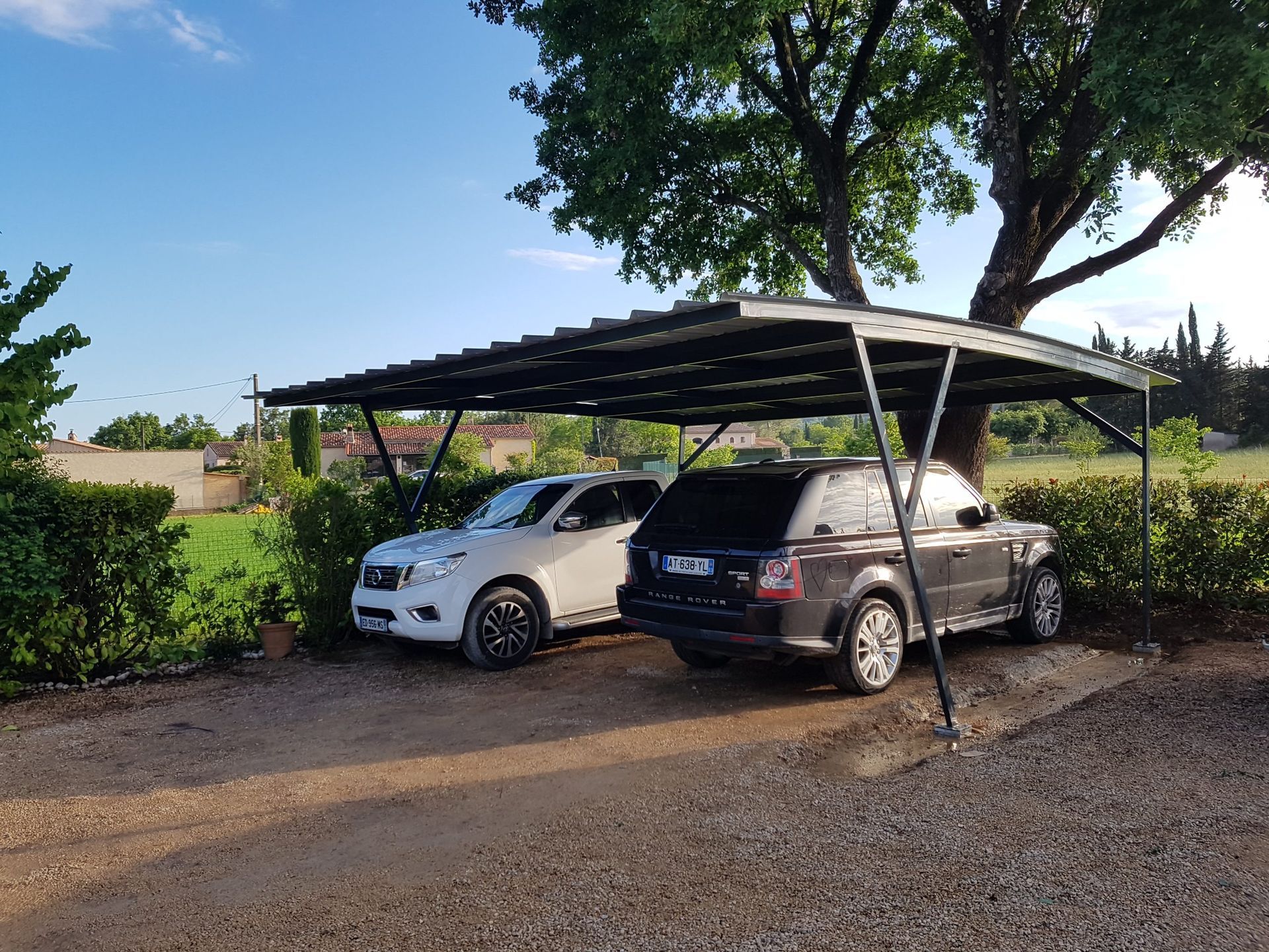 Deux voitures garées sous un abri métallique sur une allée de gravier, bordée de haies et d'arbres.