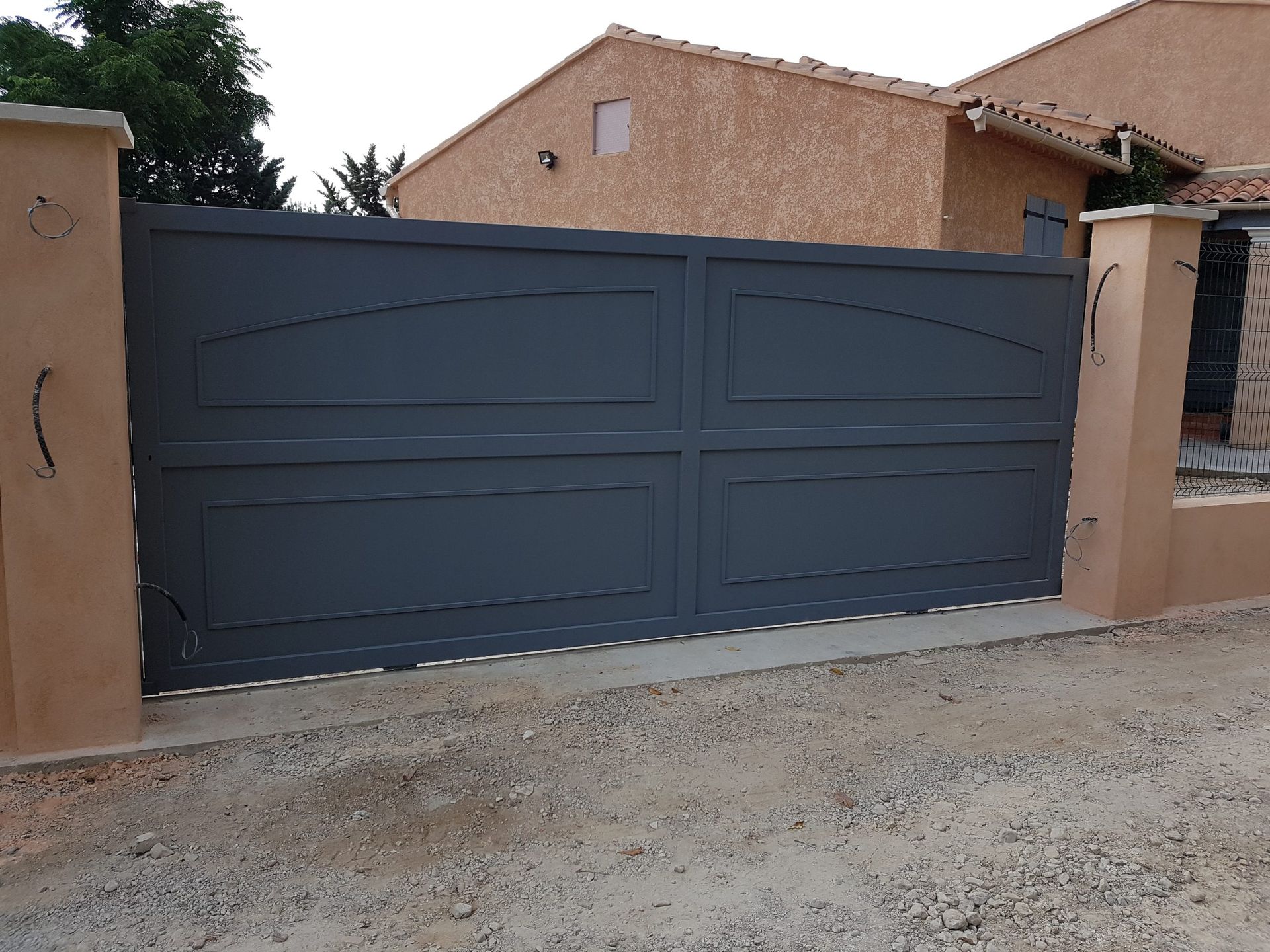 Portail d'allée en métal bleu foncé sur un mur beige devant une maison, au bord d'un chemin de gravier.